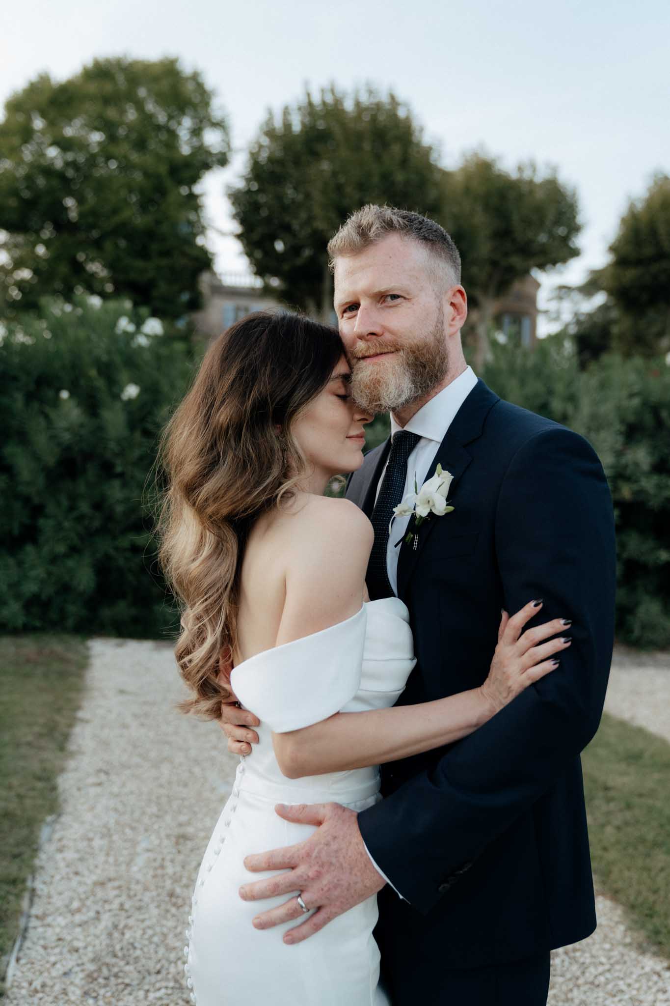 A couple portrait taken outdoors on a gravel path, with a stone building partially visible among trees in the background. The bride wears an off-the-shoulder fitted white gown with button-back detailing and has long wavy brunette hair; she rests her face close to the groom's cheek with her eyes closed. The groom wears a navy suit with a dark tie and a white floral boutonniere, and looks directly at the camera. The styling is modern and minimal. Medium portrait composition with a shallow depth of field blurring the background.