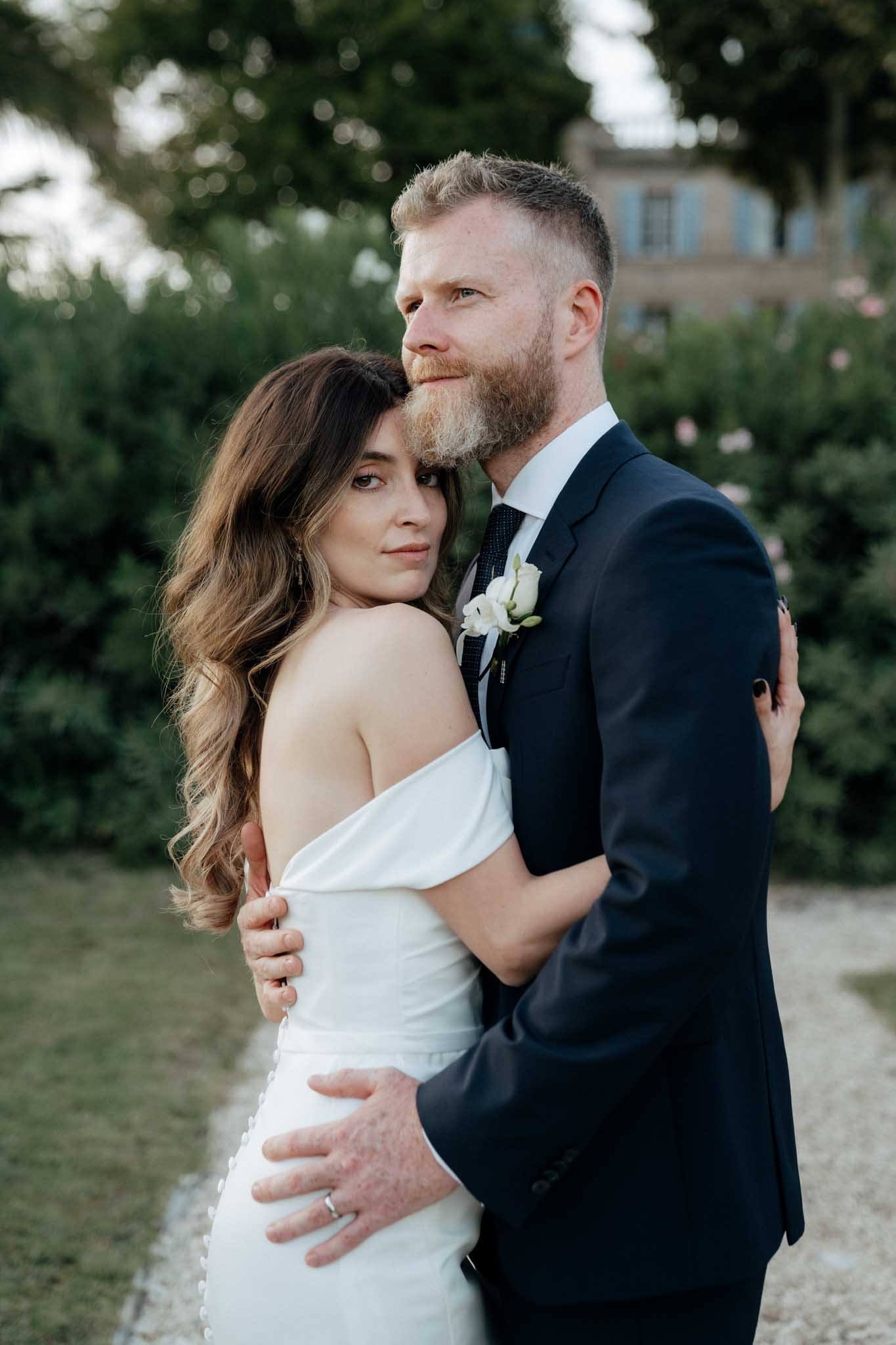 A close-up portrait of a bride and groom posing together outdoors in a garden setting, with a French chateau building with blue shutters softly visible in the background. The bride wears an off-the-shoulder white fitted gown with button detailing along the back, her long brunette hair styled in loose waves; she rests her head against the groom's cheek while looking directly at the camera. The groom wears a navy suit with a white dress shirt, navy dotted tie, and a white rose boutonniere; he holds the bride around the waist and looks slightly off-camera. The image is shot with a shallow depth of field, keeping the couple sharp against a blurred green garden backdrop.