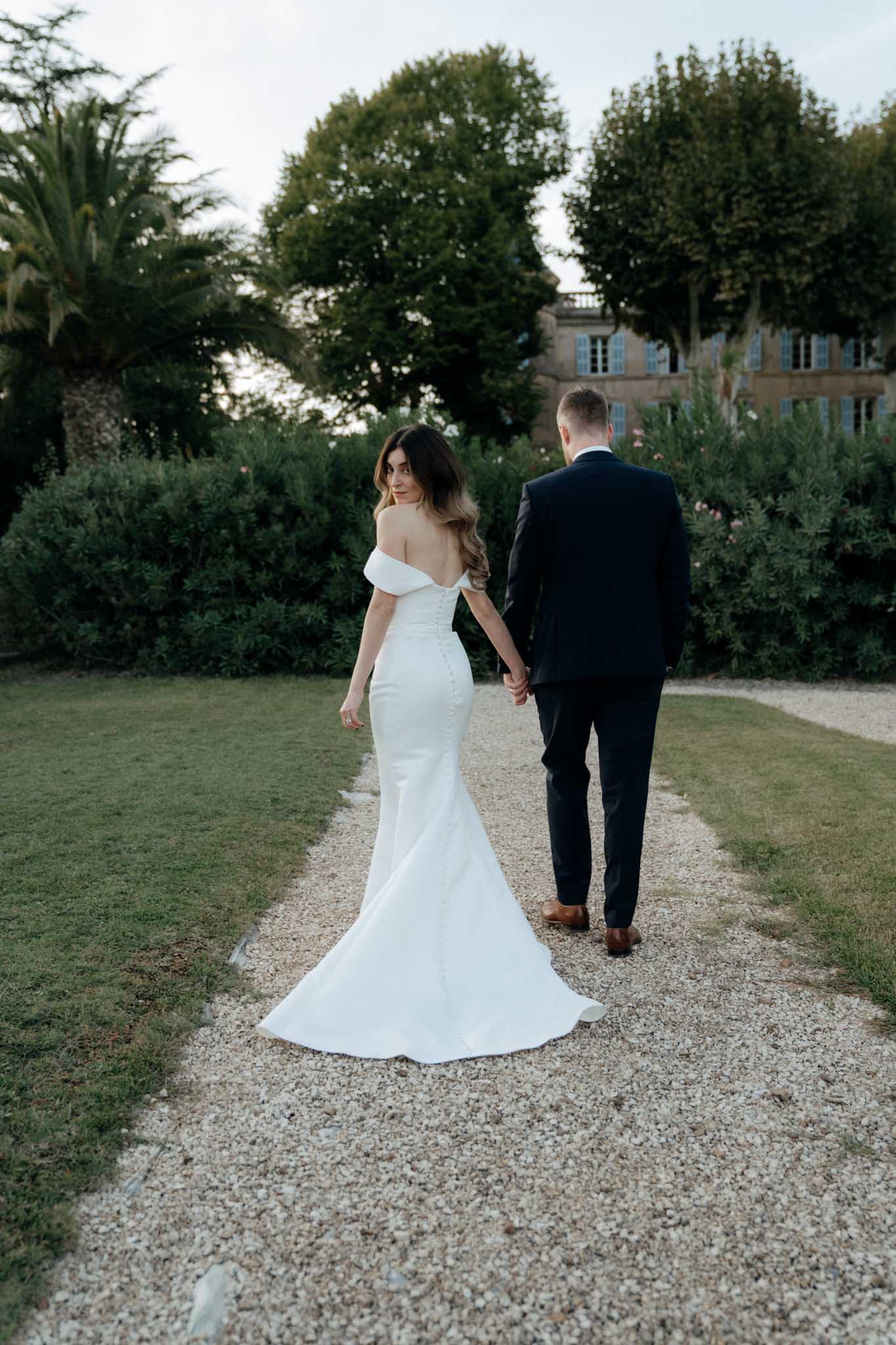 A couple portrait taken outdoors in the formal garden of a French chateau, with the bride and groom walking away from the camera along a gravel path. The bride glances back over her shoulder toward the camera, wearing a white off-the-shoulder fitted mermaid gown with a low open back, button-detail spine, and a trailing train; the groom wears a dark navy suit with tan leather shoes and holds her hand. The chateau building with pale stone facade and blue shuttered windows is visible in the background. The shot is a full-length portrait with a classic, clean styling aesthetic.