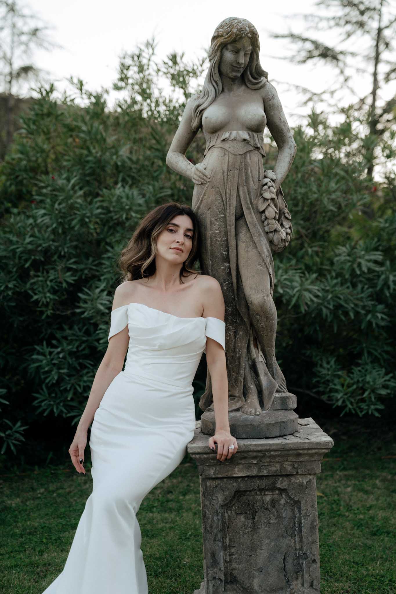 A bridal portrait taken outdoors in a formal garden setting, with the bride leaning against the stone pedestal of a classical figurative statue. The bride wears a fitted off-the-shoulder white gown with a clean, minimalist silhouette and a slight train. Her dark hair is worn in loose waves and she wears a solitaire ring. The weathered stone statue depicts a female figure holding a garland. The styling is modern and clean against the classical garden decor. Medium portrait shot, slightly low angle.