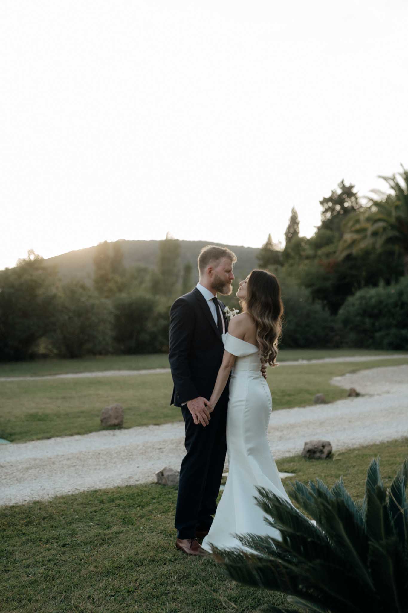 A couple portrait taken outdoors during golden hour, with warm backlight coming from behind the treeline. The groom wears a dark navy suit with a black tie and a small white boutonniere, while the bride wears a fitted off-the-shoulder white satin gown with button-back detailing and long wavy dark hair. They stand face to face holding hands on a manicured lawn, with a gravel path winding through the grounds behind them. The composition is a medium full-length portrait with a modern, minimal styling aesthetic.