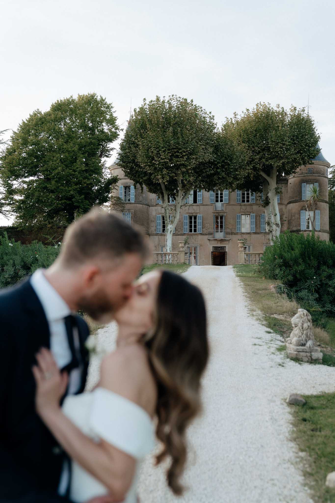 A couple shares a kiss in the foreground during an outdoor couple portrait session, intentionally photographed out of focus to draw attention to the venue behind them. The bride wears a white off-the-shoulder dress and has long wavy dark hair, while the groom is dressed in a dark navy suit with a white boutonniere. The sharp background reveals a multi-story French chateau with warm stone facade, pale blue shutters, a rounded turret, and a grand entrance framed by a balustrade, accessed via a gravel driveway flanked by stone lion sculptures. The composition uses a wide portrait orientation with the couple blurred in the foreground and the chateau in full focus. Potential venue feature image.