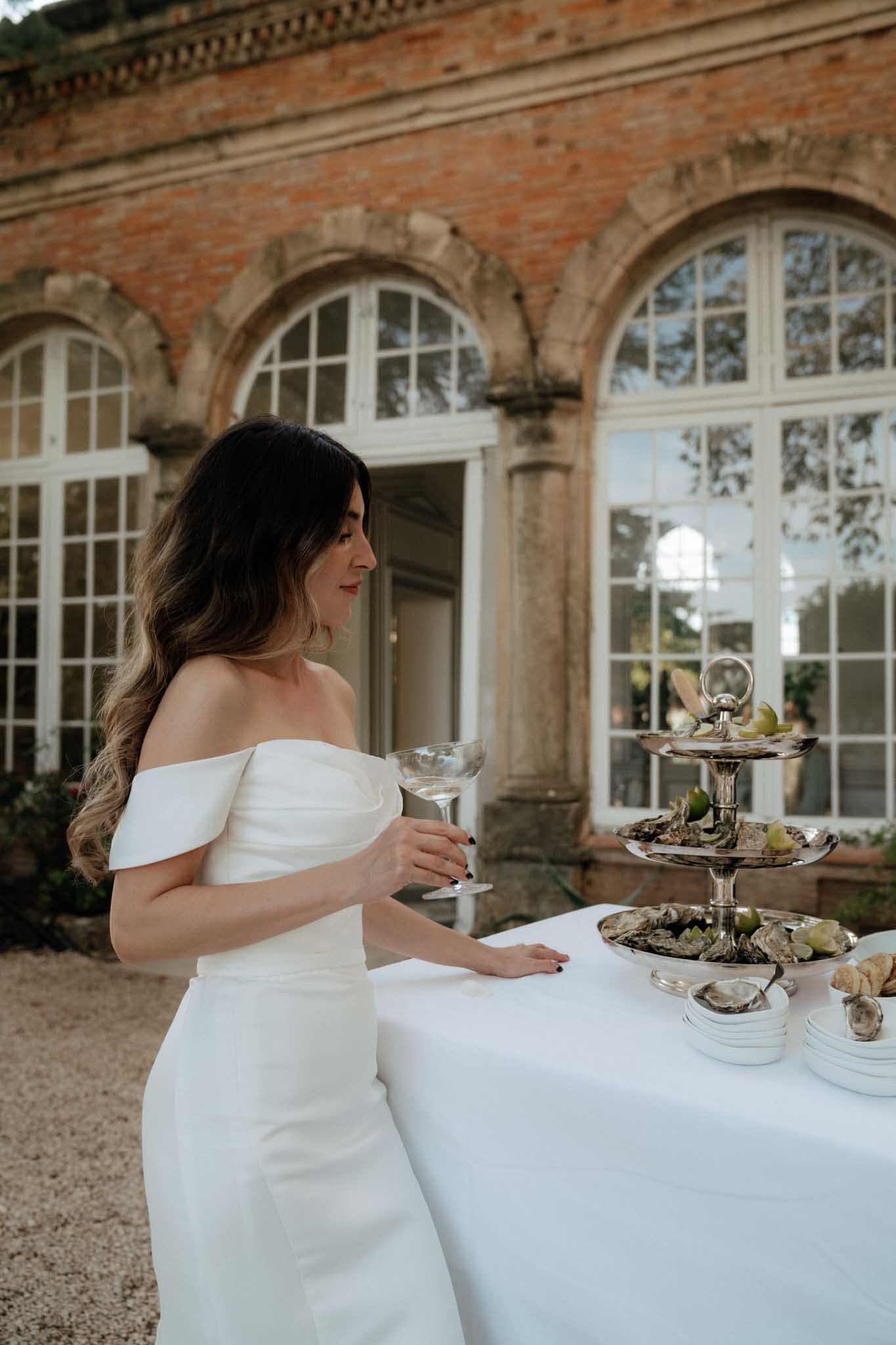 A bride stands at a cocktail hour food station outdoors in front of a red brick building with large arched white-framed windows, likely a chateau or manor orangery. She is wearing a white off-the-shoulder fitted gown with a structured fold detail at the neckline, and holds a coupe-style cocktail glass. On the white linen-covered table beside her sits a silver three-tiered seafood tower loaded with oysters and limes, accompanied by small white condiment dishes and bread. The overall styling is modern and minimal. Portrait shot from a three-quarter angle.