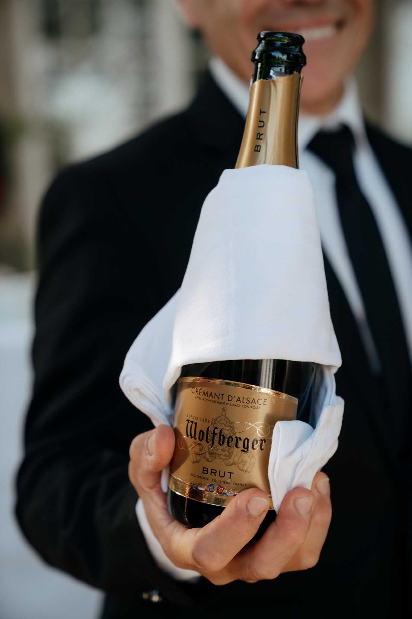 Close-up detail shot of a smiling waiter or server in a black suit and white shirt presenting a bottle of Wolfberger Crémant d'Alsace Brut toward the camera, wrapped in a white service cloth. The gold-labeled bottle is held at the forefront of the frame in sharp focus, with the server's face softly blurred in the background. This shot captures a cocktail hour or reception service moment, with the Alsatian sparkling wine choice reflecting a classic French wedding beverage selection.