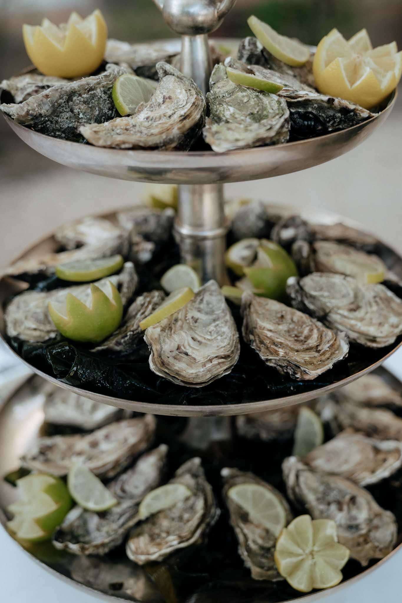 Close-up detail shot of a three-tiered silver seafood tower loaded with fresh oysters on the half shell, arranged on beds of dark seaweed across all three tiers. Each tier is garnished with lime and lemon wedges, some cut into decorative flower shapes. The display is a classic French cocktail hour or reception catering element, presented on a polished silver stand. The background is softly blurred, keeping full focus on the food presentation.