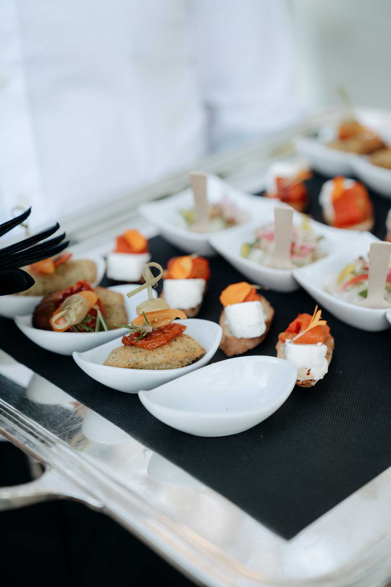 Close-up detail shot of cocktail hour canapés arranged on a silver serving tray lined with a black mat. The canapés are presented in small white teardrop-shaped individual dishes and include varieties such as blinis topped with smoked salmon and cream cheese, mini toasts with confit tomatoes and rosemary, bites with mozzarella and roasted orange vegetables, and small spoons of what appears to be a tartare or ceviche garnished with small white label picks. Black serving tongs are visible at the left edge of the tray. The presentation style is modern and refined, with a consistent white dish format across all portions.