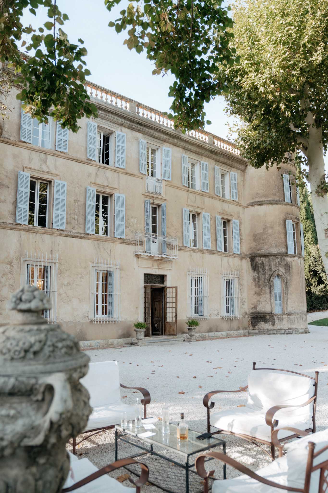 A wide exterior shot of a three-story French chateau with pale sand-colored stone facades and powder blue shutters across all windows, featuring a rounded tower section on the right side and a central entrance with ornate ironwork doors and a wrought-iron balcony above. In the foreground, a cocktail lounge area has been set up on a gravel courtyard with white-cushioned dark wood armchairs arranged around a glass-and-iron coffee table holding glassware and a bottle. A weathered stone urn sculpture is visible in the lower left corner. The overall decor palette is white and dark bronze-toned metal against the neutral stone architecture, suggesting a classic, refined outdoor cocktail hour setup. Potential venue feature image.