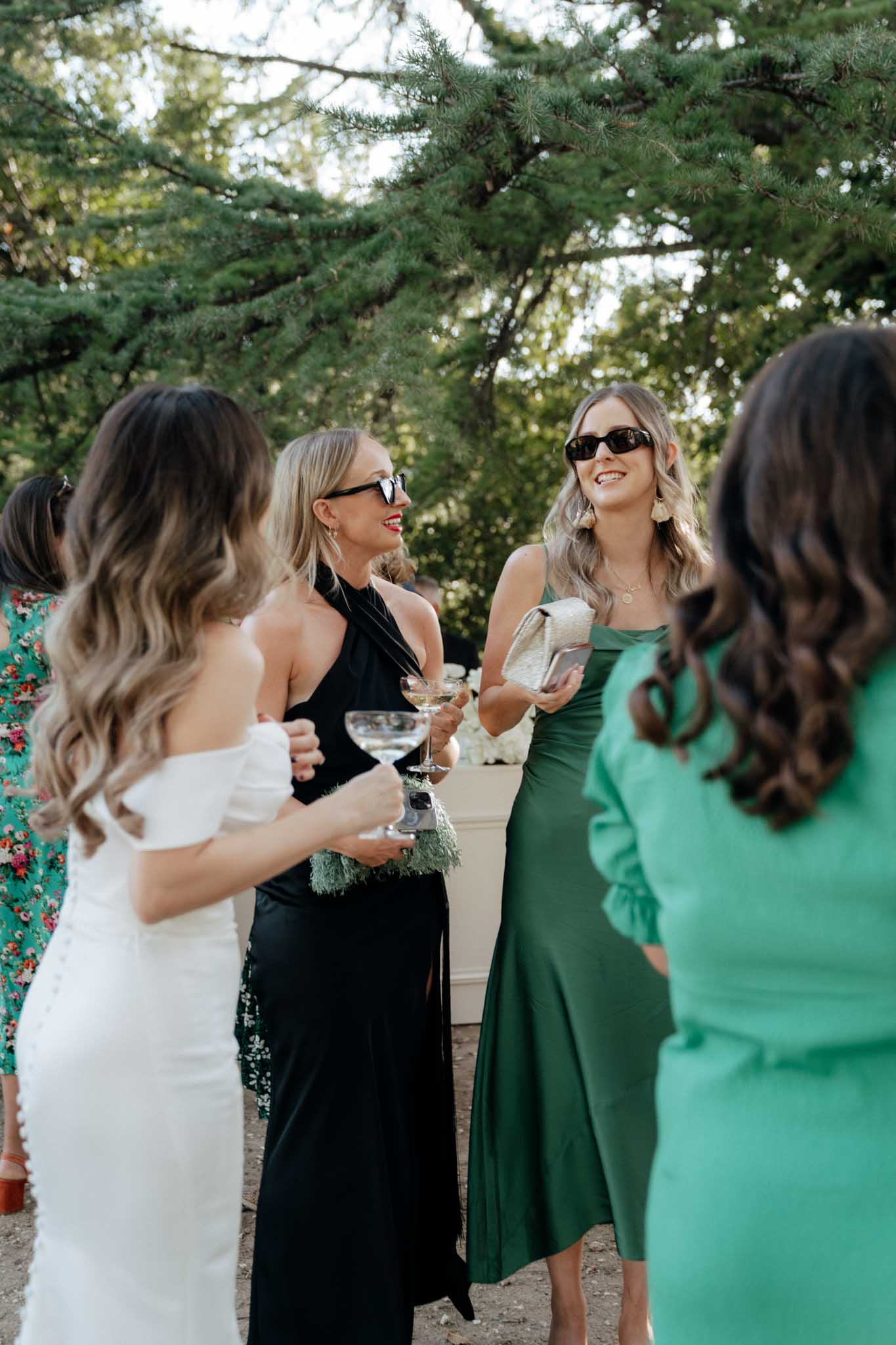 An outdoor cocktail hour scene with a small group of five women gathered in conversation, with the bride visible from behind on the left in a fitted white off-the-shoulder gown with lace detailing. The guests are dressed in a mix of a black halter-neck maxi dress, a forest green satin midi dress, a bright emerald green long-sleeve dress, and a floral print dress partially visible at the edge of the frame. Two of the guests wear black sunglasses and hold coupe glasses, likely containing cocktails. The setting appears to be a gravel terrace or courtyard with tall pine trees in the background. The overall guest styling palette leans toward deep and bright greens contrasted with black, giving the group a modern, polished look. The shot is a candid medium portrait taken at eye level.