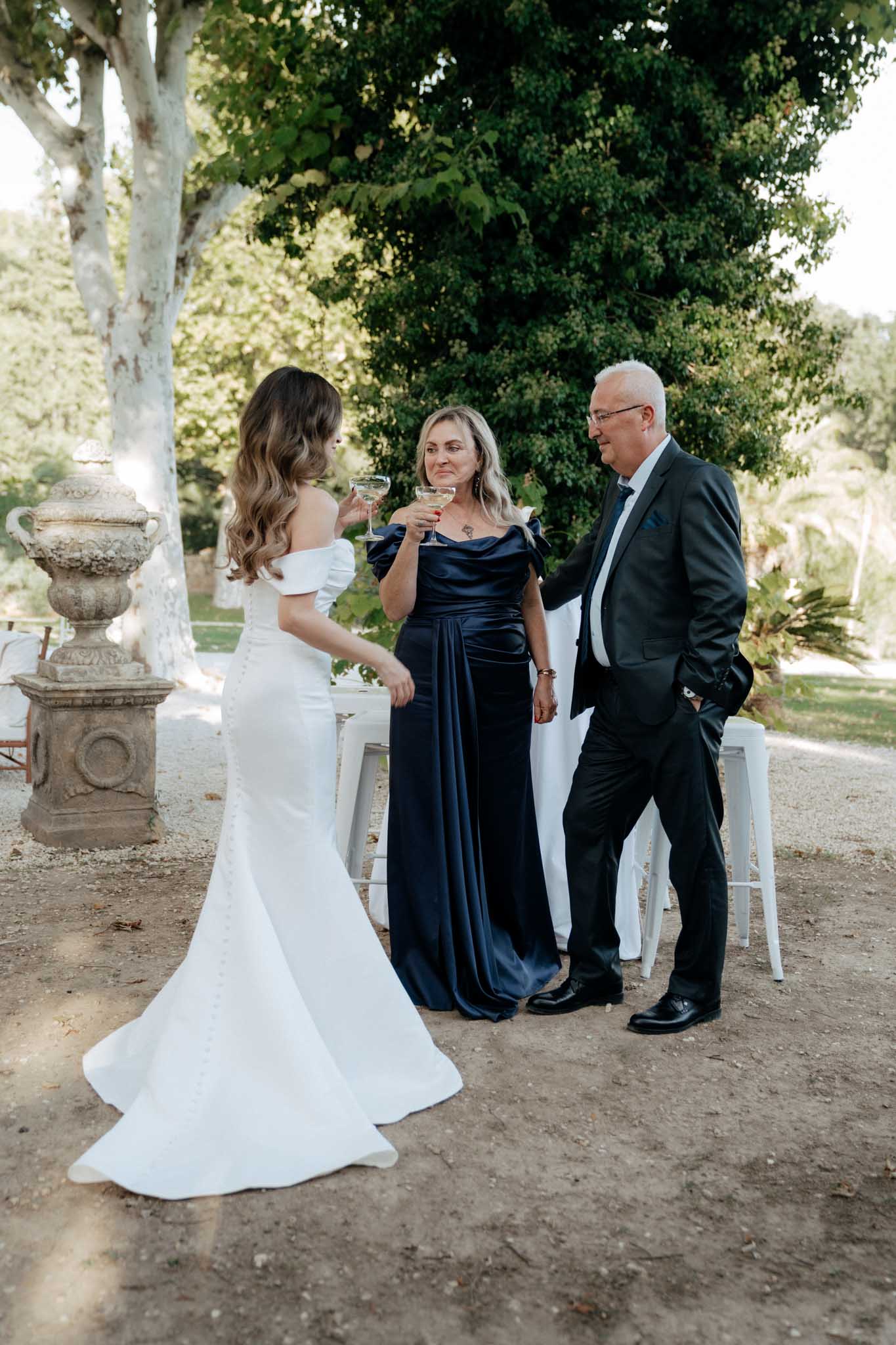 An outdoor cocktail hour scene showing the bride toasting with two guests, likely family members, in a garden setting. The bride is wearing a fitted off-the-shoulder white gown with a small train and button detail down the back, photographed from behind. The female guest is wearing a navy blue satin off-the-shoulder floor-length dress, and the male guest is in a dark suit with a tie; all three are holding coupe-style champagne glasses. A white linen-covered cocktail table and white metal bar stools are visible in the background, alongside a large ornamental stone urn planter, suggesting a classic French estate or chateau setting. The shot is a medium full-length portrait taken from a slight distance.