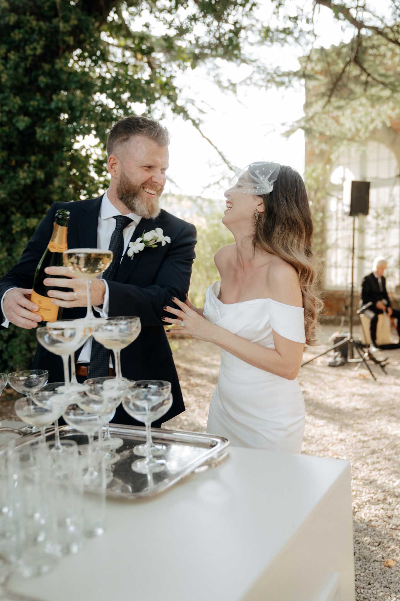 The bride and groom are pouring champagne into a coupe glass tower during an outdoor cocktail hour or reception, set on a gravel terrace with a classical stone building visible in the background. The groom wears a navy suit with a dark tie and a white orchid boutonniere, and is holding a bottle of Veuve Clicquot champagne; the couple is laughing and looking at each other. The bride wears an off-the-shoulder fitted white gown with a draped bodice and a small polka-dot birdcage veil pinned in her long wavy hair. The champagne tower is arranged on a white bar surface with a silver tray, and a musician can be seen seated in the background, suggesting live music at the event. The shot is a medium portrait capturing both figures from roughly waist height, with a candid, joyful feel.