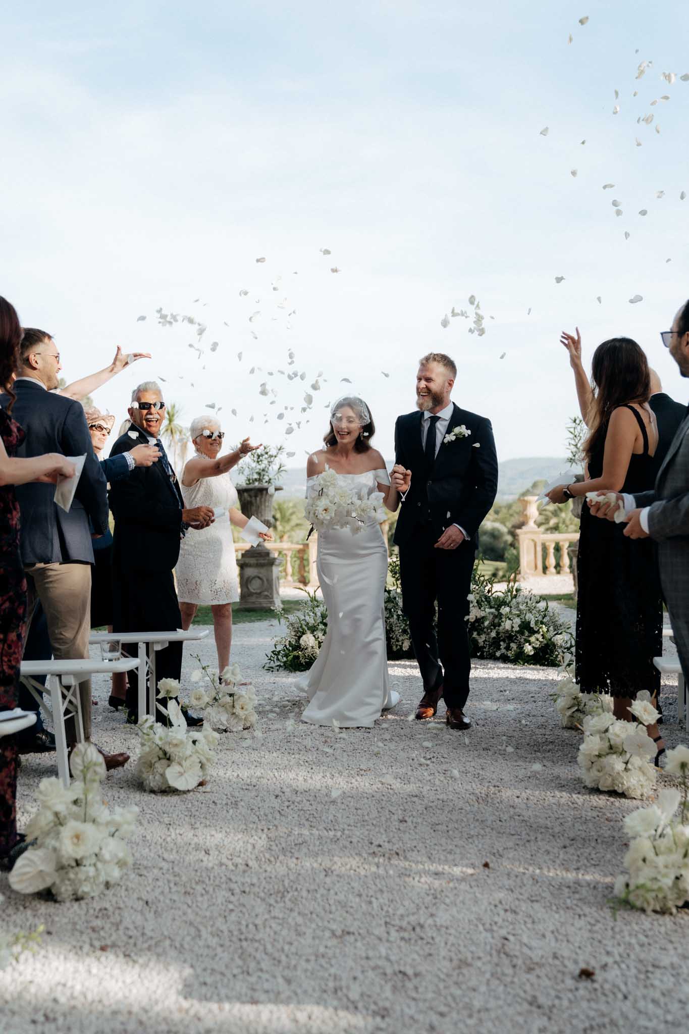 The bride and groom walk back down the aisle together following an outdoor ceremony, as approximately ten guests on either side toss white flower petals into the air around them. The ceremony took place on a gravel terrace with a classic balustrade visible in the background, consistent with a French chateau or villa setting. The bride wears a fitted off-the-shoulder ivory satin gown with a short birdcage veil and carries a loose bouquet of white peonies and ivory blooms; the groom wears a dark navy suit with a white boutonniere and grey tie. The aisle is lined with low clusters of white floral arrangements including peonies, ranunculus, and greenery, and white folding chairs are visible on both sides; the overall decor palette is all-white with a classic, clean aesthetic. This is a medium wide shot taken straight down the aisle.