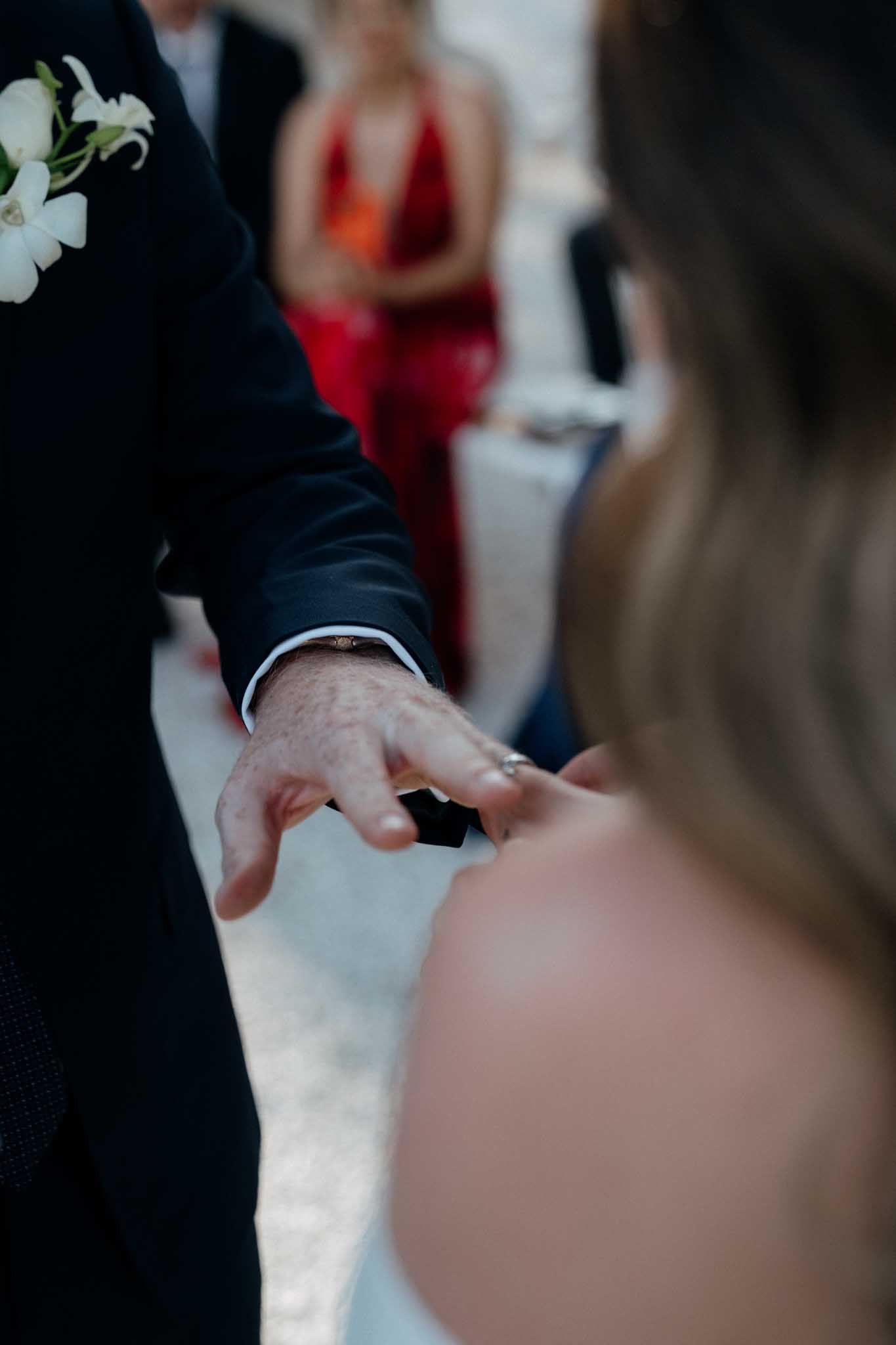 Close-up detail shot of a ring exchange moment during an outdoor wedding ceremony. The groom, wearing a navy or black suit jacket with a white shirt cuff visible and a white orchid boutonniere, is placing a ring onto the bride's finger, who is shown from behind in a strapless white dress. A small dark ring box is visible in the groom's hand. In the soft-focus background, at least one guest in a red floor-length dress is visible watching the ceremony.