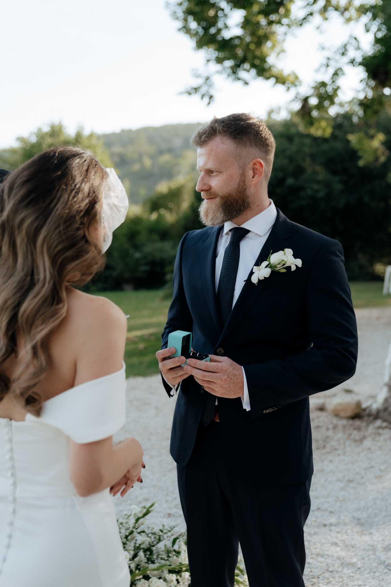 The groom holds an open light turquoise ring box toward the bride during what appears to be an outdoor ceremony exchange of rings on a gravel path. The groom wears a navy suit with a dark knit tie, white shirt, and a white floral boutonniere featuring small roses or ranunculus. The bride, seen from behind over the shoulder, wears an off-the-shoulder white gown with a button-back detail and has wavy brunette hair styled with a white bow or ribbon hair accessory. The shot is a close-up portrait framing just the two of them, with white florals visible at ground level along the ceremony path.