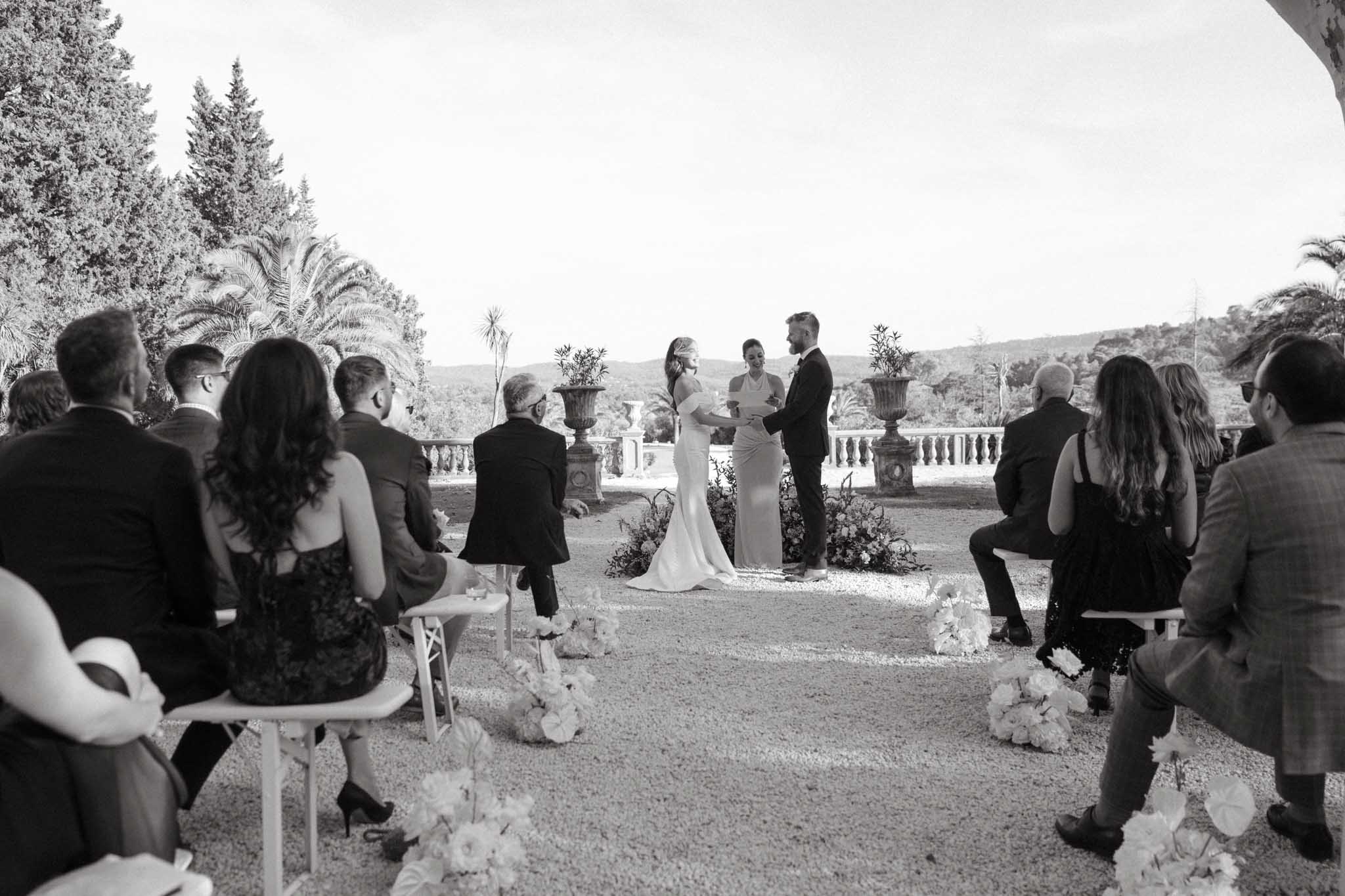 This black-and-white image shows an outdoor wedding ceremony taking place on a gravel terrace of what appears to be a French villa or chateau, with a stone balustrade, ornamental urns, and a panoramic view of hills in the background. The bride, wearing a sleek fitted gown, and the groom, in a dark suit, stand facing each other holding hands while an officiant addresses them, with low floral arrangements at ground level flanking the ceremony space. Approximately 12–15 guests are seated on white folding chairs arranged in two rows along an aisle, dressed in formal attire. The aisle is lined with clusters of round blooms placed directly on the gravel, and the overall styling is classic and refined with a strong contrast between the bright sky and dark foliage in the image tones. Wide shot taken from beneath a covered archway or portico looking toward the altar.