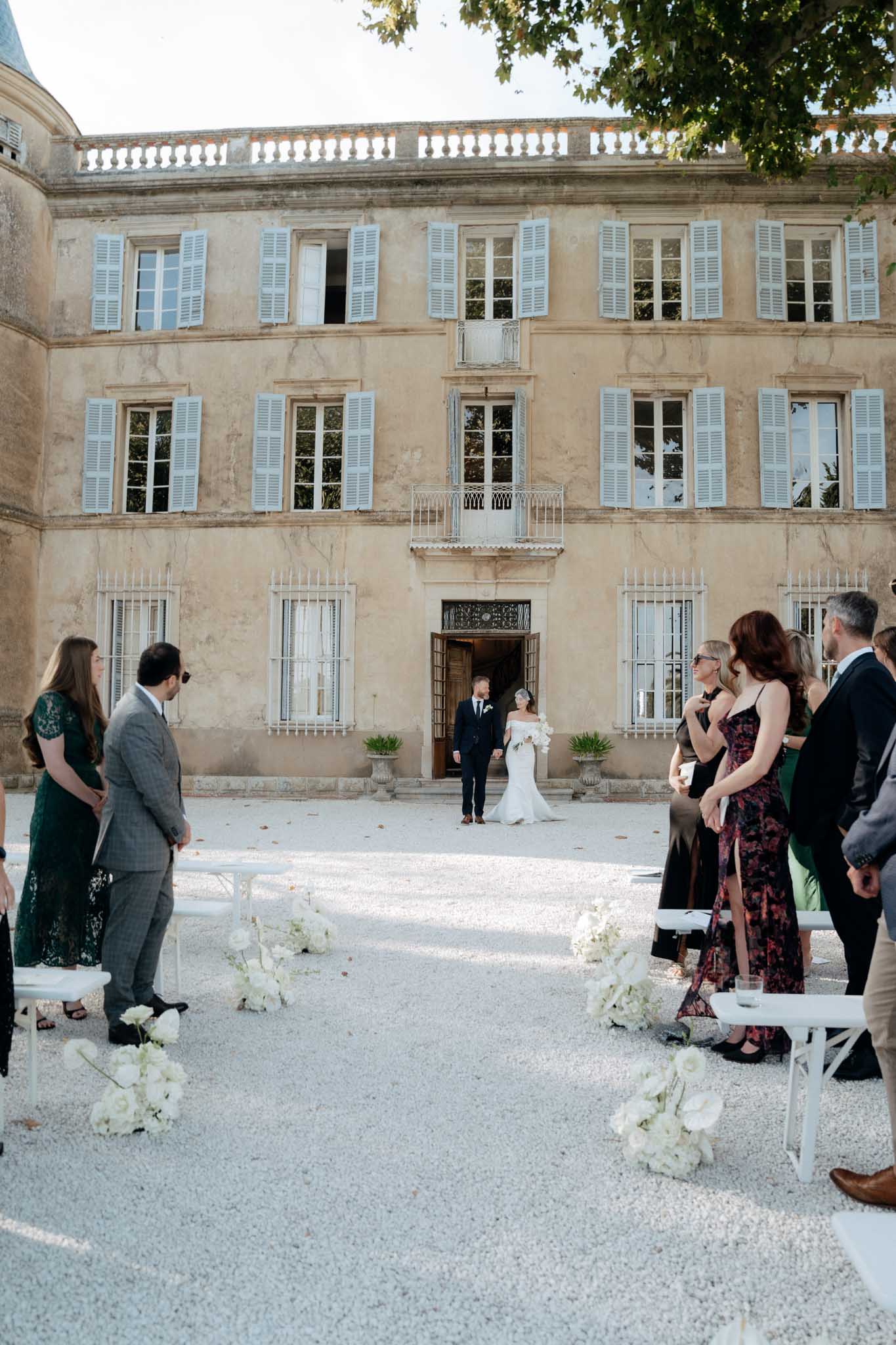 An outdoor wedding ceremony processional taking place on a gravel courtyard in front of a three-story French chateau with pale blue shutters and ornate ironwork balconies. The bride, wearing an off-the-shoulder fitted white gown and carrying a white bouquet, walks alongside the groom in a dark navy suit as they emerge through the chateau's wooden entrance doors toward the aisle. Approximately 20–30 guests stand on either side of the aisle, dressed in formal attire including dark green lace, floral maxi dresses, and dark suits. The aisle is flanked by white folding chairs and low arrangements of white roses and white blooms placed at each row end, creating a clean, monochromatic floral palette that complements the classic, formal styling. The wide shot captures the full facade of the chateau as a backdrop, centering the couple at the entrance. Potential venue feature image.