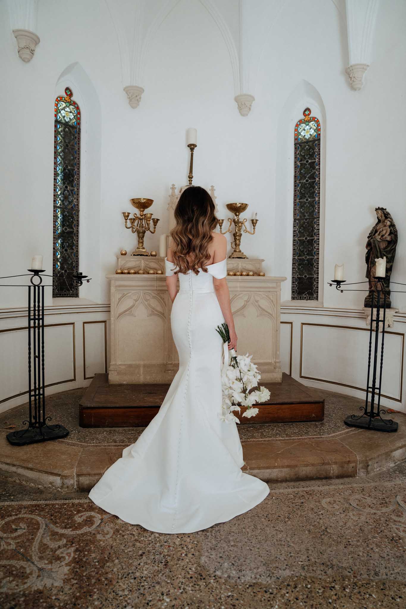 A bridal portrait taken from behind inside a small chapel or private oratory, with the bride standing at the altar. She wears a fitted off-the-shoulder white mermaid gown with a button-detail back seam and a modest train, and holds a cascading bouquet of white orchids with dark green stems. The chapel interior features white arched walls, narrow stained glass windows with red, blue, and gold detailing, a carved stone altar dressed with brass candelabras and white pillar candles, tall wrought-iron floor candleholders, and a painted religious statue to the right. The floor is a patterned terrazzo. The overall styling is clean and modern against a classic religious setting. Full-length portrait shot from a medium distance.