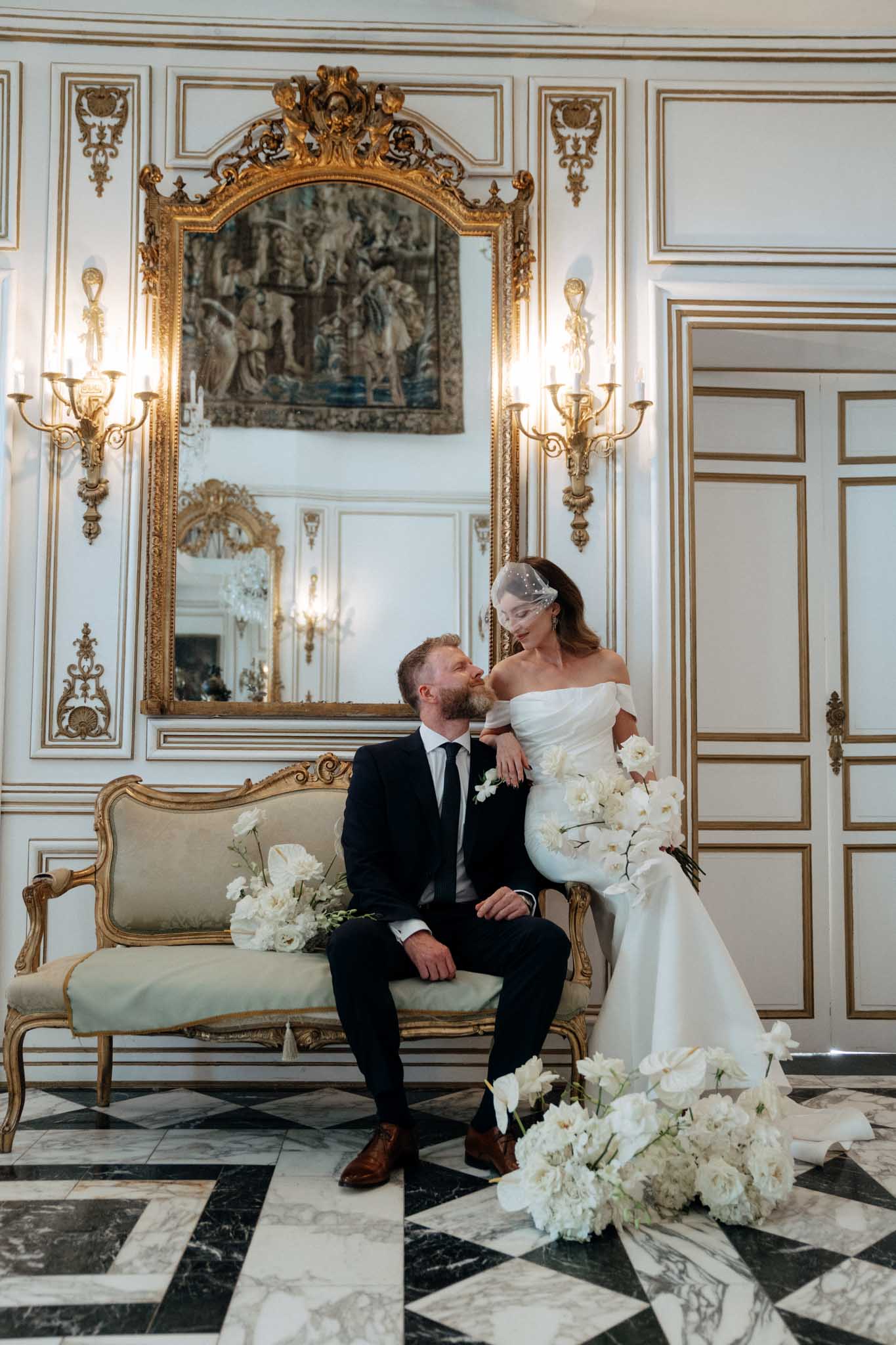 A couple portrait taken indoors in a grand French interior featuring white-and-gold boiserie paneling, gold-framed wall sconces with candle-style lighting, a large ornate gold mirror with a tapestry reflected within it, and a black-and-white marble diamond-patterned floor. The groom, wearing a dark navy suit with a black tie and brown leather oxford shoes, is seated on a gilded Louis XVI-style sofa upholstered in pale sage green, looking up at the bride. The bride wears an off-the-shoulder fitted white gown with a short birdcage veil adorned with small pearls or crystals, and she leans against the groom while looking down at him. Multiple arrangements of white flowers — including phalaenopsis orchids, garden roses, ranunculus, and sweet peas — are placed on the sofa beside the groom and arranged on the marble floor in the foreground, creating an all-white floral installation that frames the couple. The overall styling is classic and formal with a monochromatic white floral palette. Wide portrait shot.