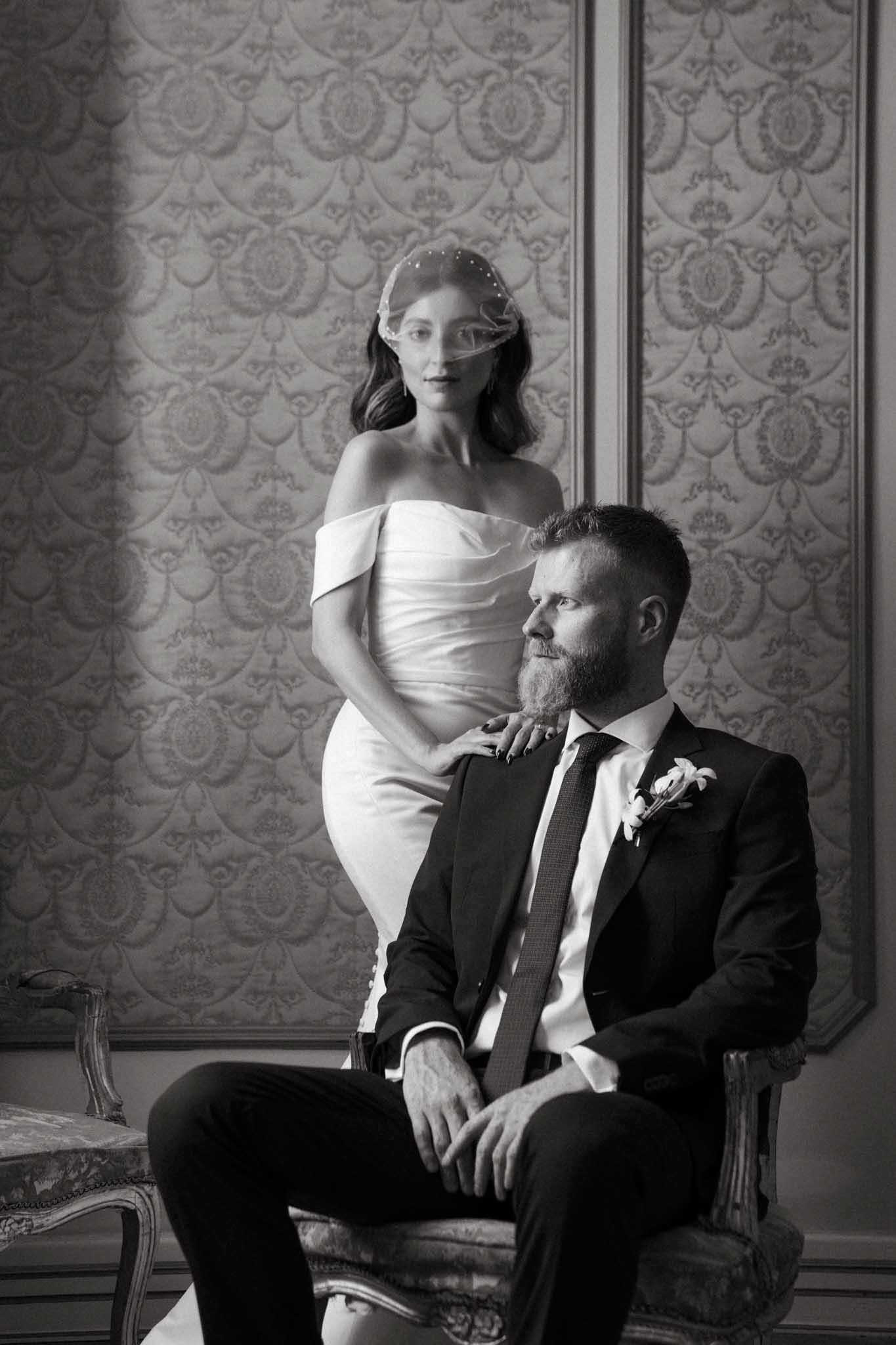 A black-and-white couple portrait taken indoors against an ornate damask-patterned wall, likely inside a chateau or formal reception venue. The bride stands behind the seated groom, wearing a short off-the-shoulder fitted dress and a birdcage veil positioned across her face, with her hand resting on his shoulder. The groom sits in an antique upholstered chair wearing a dark suit, textured tie, and a small floral boutonniere, gazing to one side. The image has strong contrast with deep shadows and bright highlights, giving it a classic editorial quality. The composition is a mid-length portrait with both subjects framed against the decorative wall paneling.