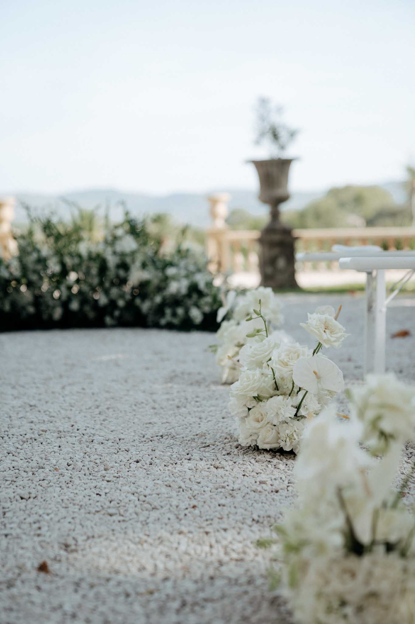 A close-up detail shot of an outdoor wedding ceremony aisle setup on a gravel surface, with clusters of all-white floral arrangements placed along the aisle edge. The bouquets consist of white garden roses, ranunculus, and lisianthus. White chairs are partially visible on the right side of the frame. In the soft-focus background, a stone urn planter sits atop a classical balustrade terrace, with a dense hedge of white flowering shrubs behind it, suggesting a formal French estate or chateau garden setting. The overall décor palette is entirely white and green, reflecting a classic, refined ceremony aesthetic.
