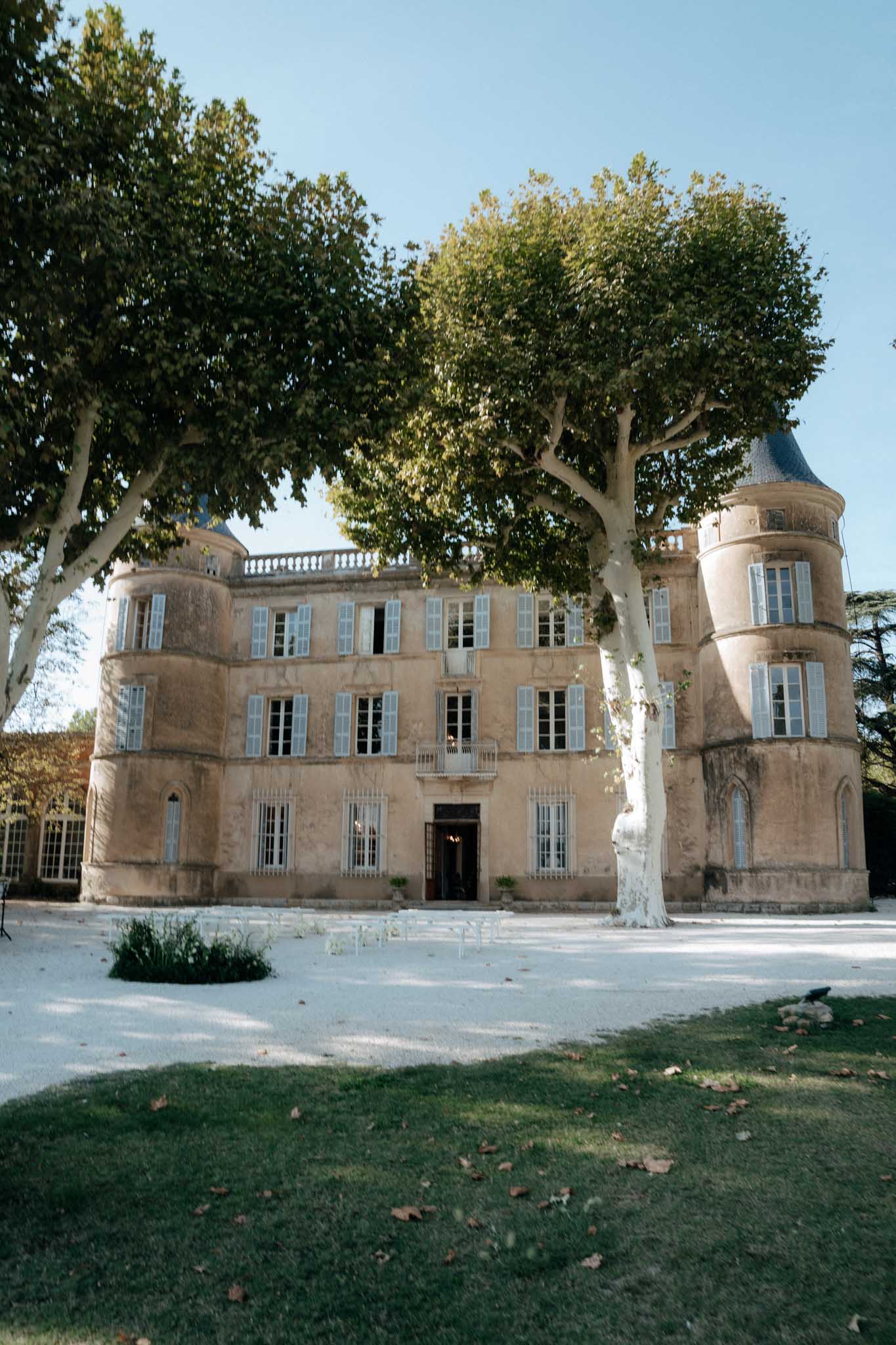 Wide exterior shot of a French chateau featuring warm honey-toned limestone facade with pale grey-blue shutters, a central entrance with an ornate iron-railed balcony above, and two rounded corner towers with slate-grey conical roofs. The three-storey building is framed by large plane trees with white-grey bark in the foreground, and the grounds include a pale gravel courtyard with rows of simple white ceremony chairs set up facing the entrance. A greenhouse or orangery wing is partially visible on the left side of the building. Potential venue feature image.