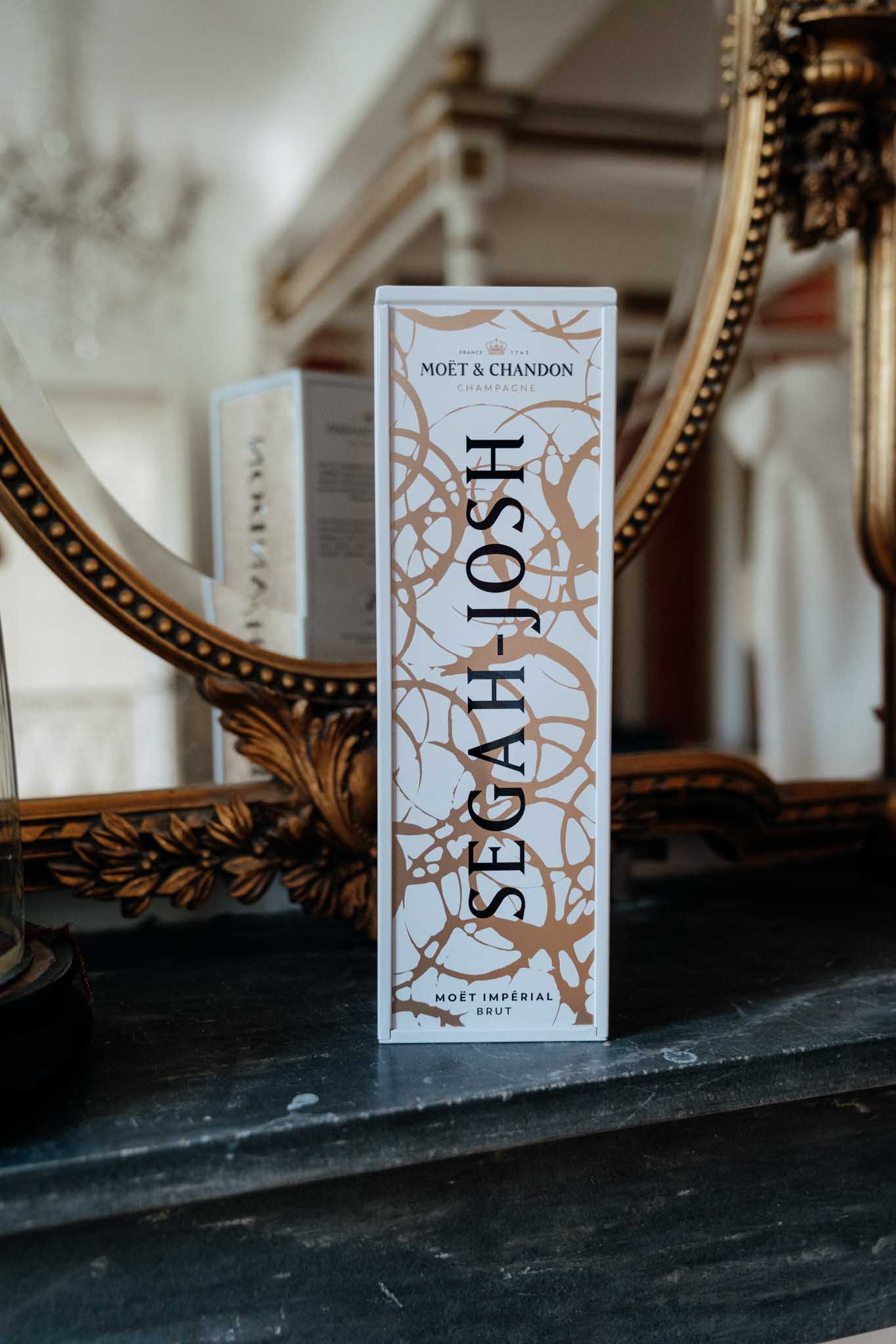 Close-up detail shot of a personalized Moët & Chandon Impérial Brut champagne gift box customized with the couple's names 'Segah & Josh' in large black lettering over a white and copper/rose-gold abstract pattern. The box is placed on a dark marble mantelpiece in front of a large ornate gold-framed mirror, with a blurred interior room visible in the reflection. The styling reflects a classic, formal aesthetic with the gold mirror frame complementing the warm metallic tones of the champagne box packaging.
