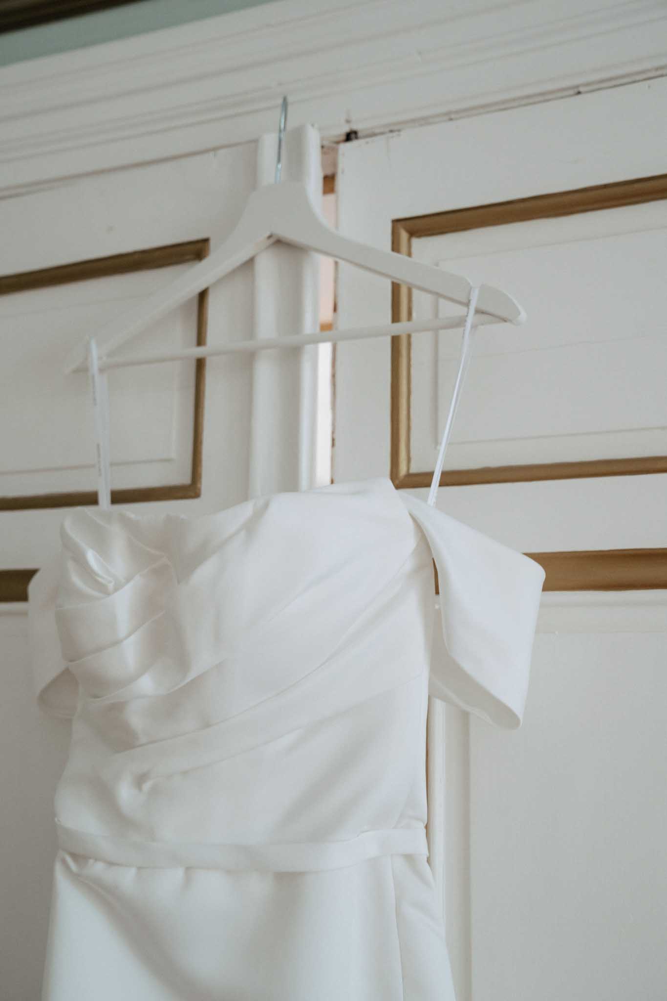 A getting-ready detail shot of an ivory satin wedding dress hanging on a white wooden hanger against an ornate white-painted door with gold-toned raised panel molding. The dress features a structured off-the-shoulder or cold-shoulder bodice with soft diagonal draping across the front and what appear to be detachable or built-in cap sleeves. The skirt is slim and minimalist in style. The composition is a close-up portrait orientation, centering the bodice of the dress with the bottom of the skirt slightly cropped.