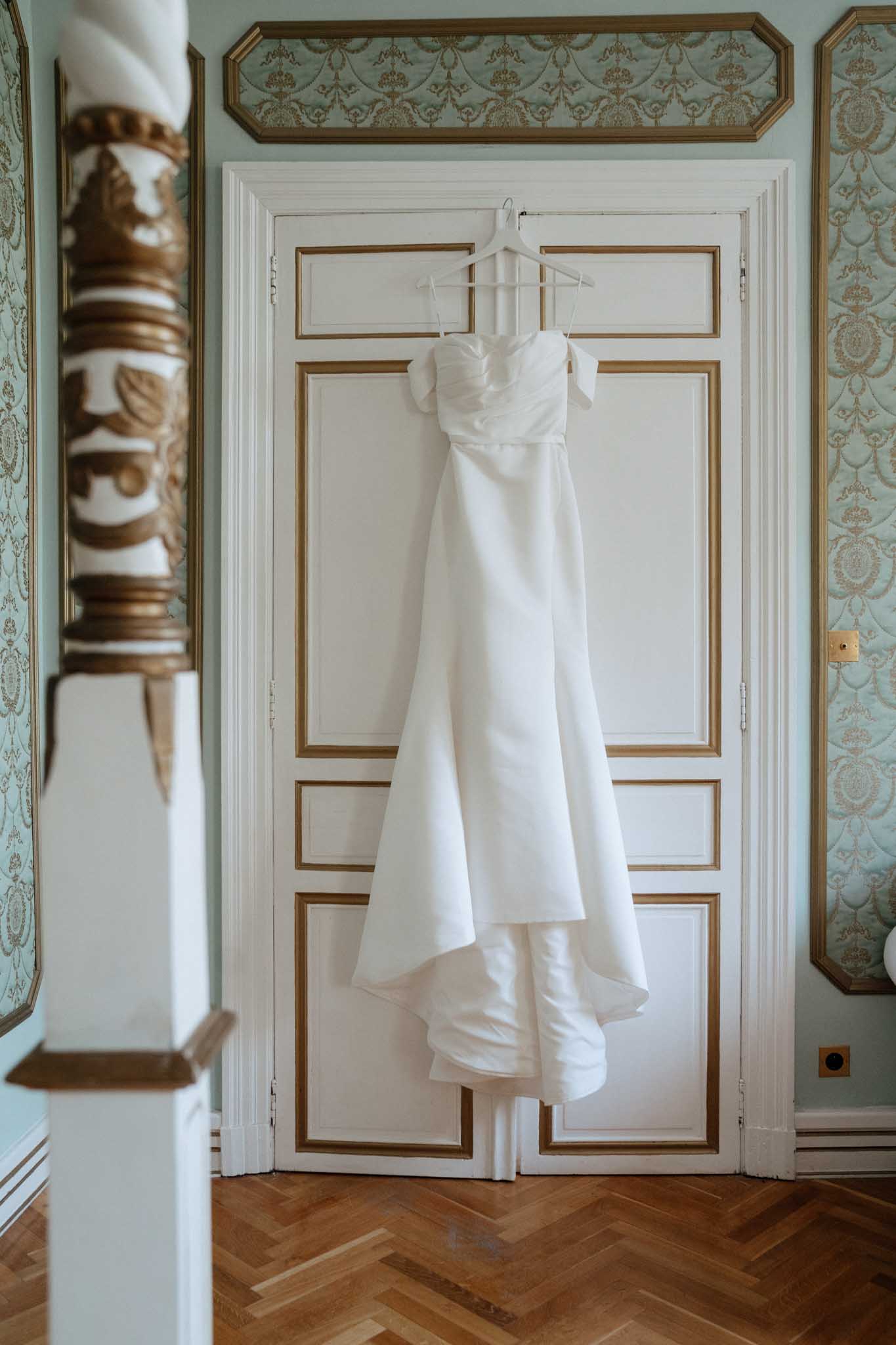 A getting-ready detail shot of a white wedding dress hanging on a white hanger from a tall double door inside a formal French interior room. The gown features an off-the-shoulder or strapless sweetheart neckline with ruched bodice detailing, a clean A-line skirt in a smooth satin fabric, and a pooling train. The door is white with gold-trimmed raised panel molding, flanked by pale sage green damask-patterned wallpaper with gold accents. A gilded decorative column or balustrade element is visible in the left foreground, and herringbone parquet flooring is visible at the base. The composition is a medium full-length shot centered on the dress against the door.