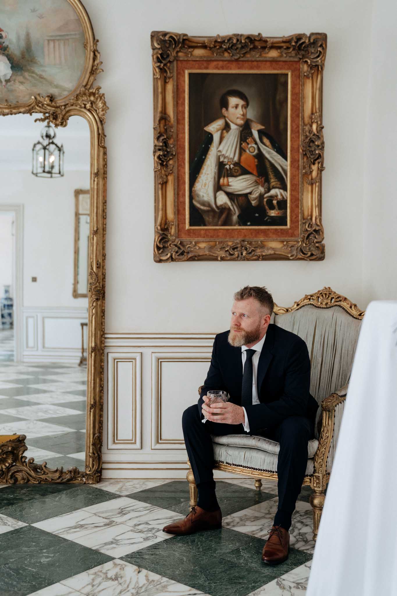 A groom sits alone in a gold-framed Louis XVI-style armchair with grey upholstery, holding a crystal whiskey glass and gazing to one side in a candid, contemplative moment. He is wearing a dark navy suit with a white dress shirt, dark tie, and brown leather Oxford shoes. The interior setting is a formal chateau salon with a green and white marble checkerboard floor, white wainscoting with gilt detailing, and a large ornate gold-framed mirror leaning against the wall. Directly above him hangs a large oil portrait painting of a nobleman in ceremonial dress within a gilt frame, creating a deliberate visual contrast between the subject and the historical figure above. The composition is a medium portrait shot taken from a slightly low angle, with natural light coming from the left.