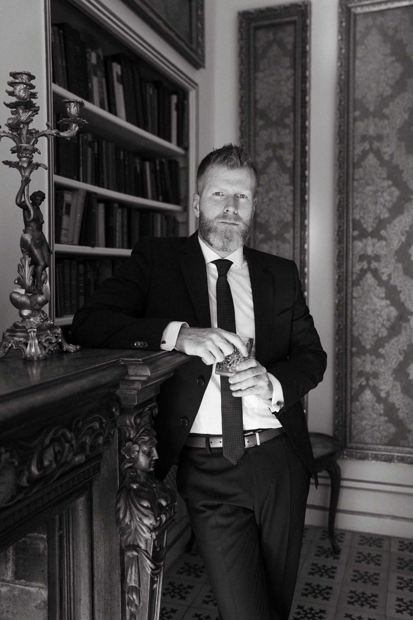 This black-and-white portrait shows a bearded groom in a dark suit, white dress shirt, and dark tie, leaning against an ornate carved fireplace mantel while holding a glass tumbler. The image features strong contrast with deep shadows in the background and crisp highlights on the subject's face and shirt. The interior setting includes built-in bookshelves filled with books, a decorative multi-arm candelabra on the mantelpiece, patterned wallpaper in ornate frames on the wall, and a geometric-tiled floor, all suggesting a classic chateau or manor house library. The composition is a medium-length portrait shot, with the groom making direct eye contact with the camera.