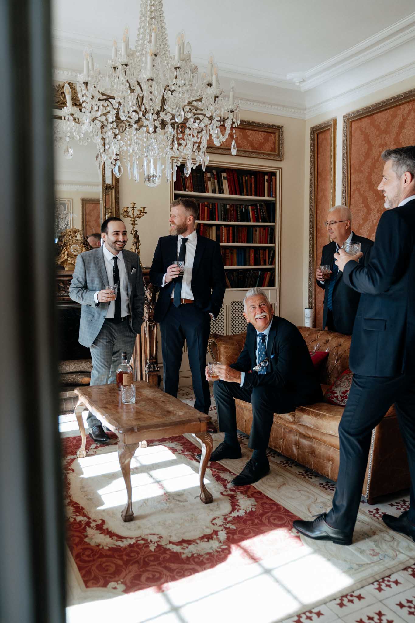 Four men are gathered in an ornate interior room, drinking whisky and conversing in a relaxed pre-ceremony getting-ready scene. The room features a large crystal chandelier, gold-framed mirrors, warm terracotta-toned wall panels, a built-in bookshelf filled with leather-bound books, and a tan leather Chesterfield sofa over a red and cream floral rug. Three of the men wear dark navy suits, while one wears a light grey checked suit with a black tie; a whisky bottle sits on a rustic wood coffee table in the foreground. The shot is taken from outside a doorway or window, giving a candid, slightly voyeuristic wide-angle perspective on the group.