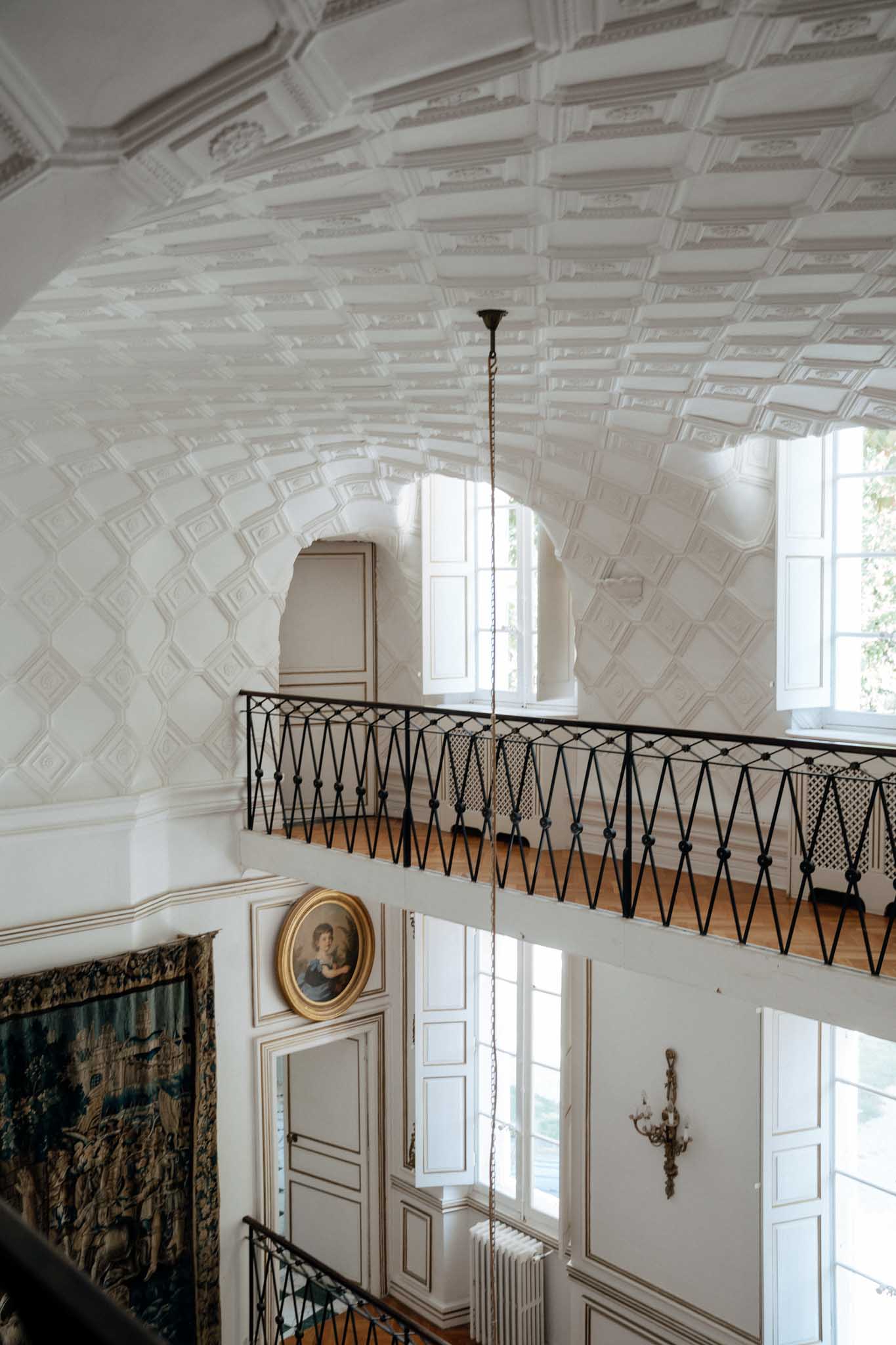 Interior architectural detail shot of a French chateau stairwell or grand hall, captured from a high angle looking across a mezzanine level. The vaulted ceiling features elaborate white plasterwork with a geometric coffered pattern, and a black wrought iron balustrade with a diamond lattice design runs along the upper gallery. The lower level walls are paneled in white with gold trim, and a large antique tapestry in teal and brown tones hangs on the left wall alongside an oval gilt-framed portrait painting. A single pendant light hangs from the ceiling on a chain, and tall French windows allow natural light to filter into the space. No people are present in the image. Potential venue feature image.
