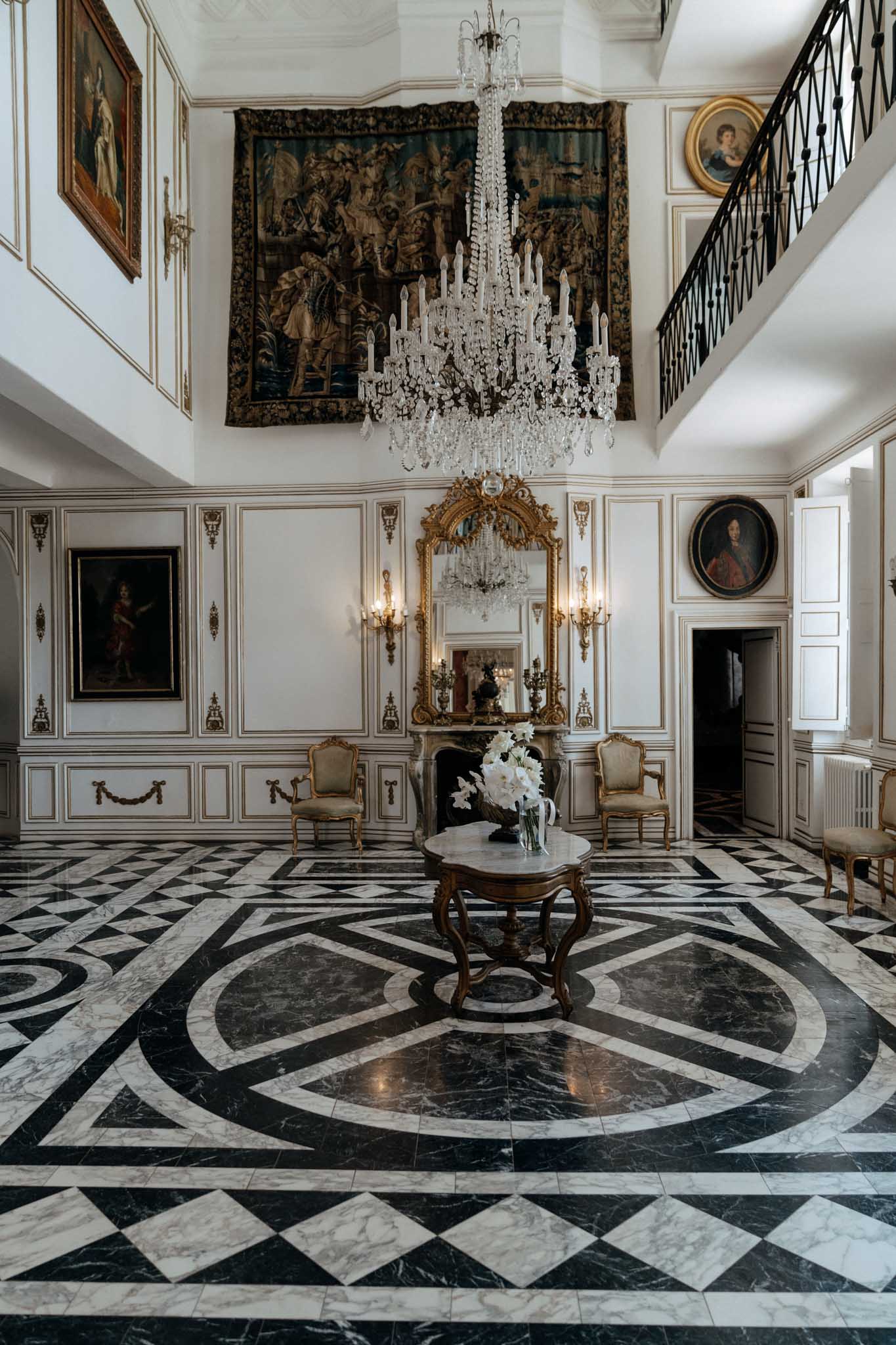 A wide interior shot of a grand chateau entrance hall or salon, with no people present. The double-height room features white paneled walls with gold gilt detailing, a large multi-tiered crystal chandelier hanging from the ceiling, and a dramatic Flemish-style tapestry mounted on the upper wall. A black and white geometric marble floor with a circular medallion pattern dominates the foreground. At center stands a carved walnut occasional table holding a vase of white amaryllis or lily blooms. A marble fireplace with an ornate gold-framed mirror and flanking gold wall sconces sits against the back wall, with two gold-framed Louis XV-style armchairs positioned on either side. Multiple oil portraits in gilded frames hang throughout the room, and a wrought-iron mezzanine balcony is visible on the upper right. The overall style is classic 18th-century French interior. Potential venue feature image.