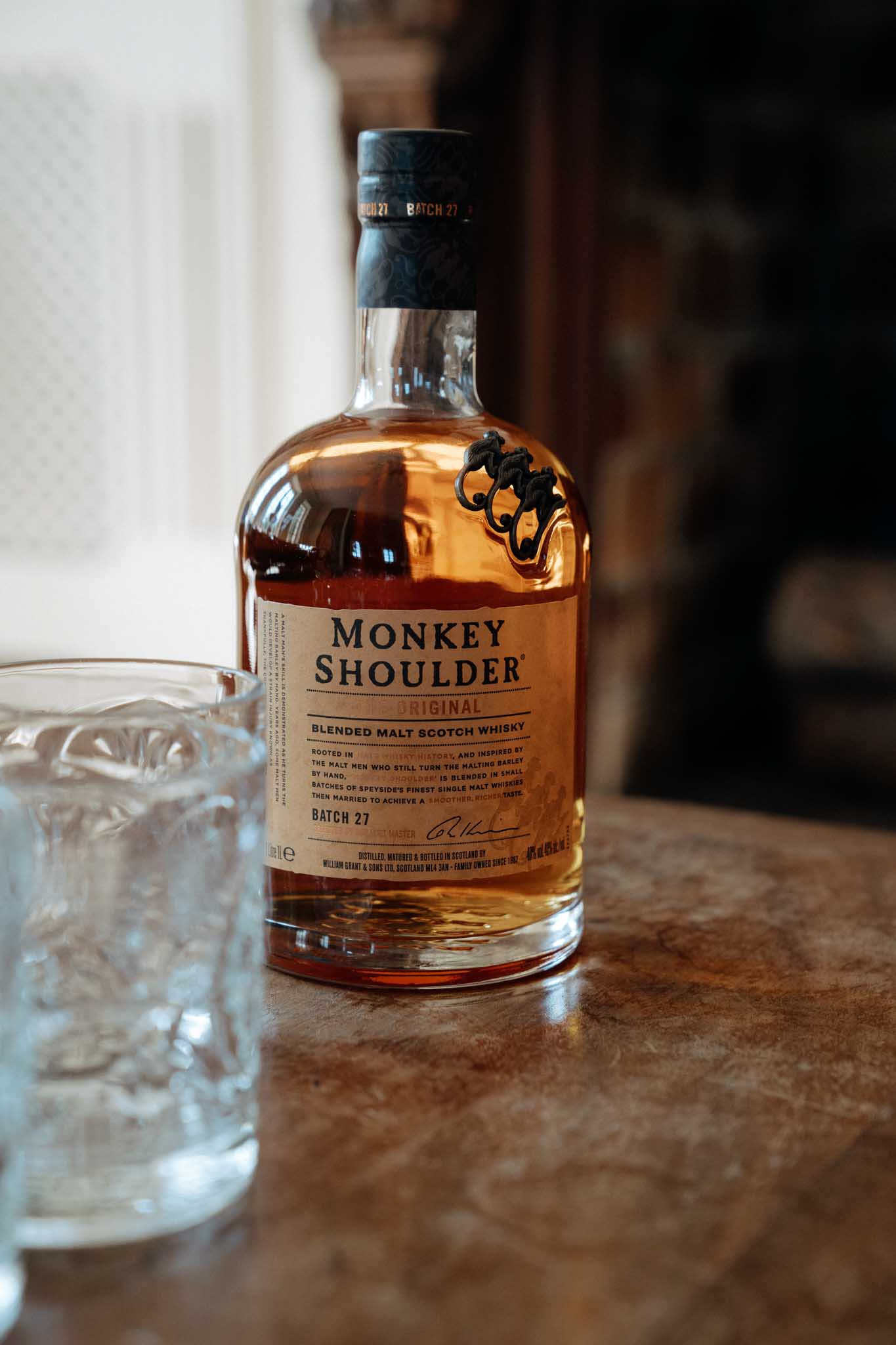 This image does not appear to be a wedding photograph. It shows a close-up detail shot of a bottle of Monkey Shoulder Original Blended Malt Scotch Whisky (Batch 27) placed on a brown surface alongside an empty cut-crystal rocks glass. The background is softly blurred with dark and light tones suggesting an indoor setting. This may have been captured as a bar or drinks detail shot, but it contains no people, wedding attire, floral arrangements, or other wedding-related elements.