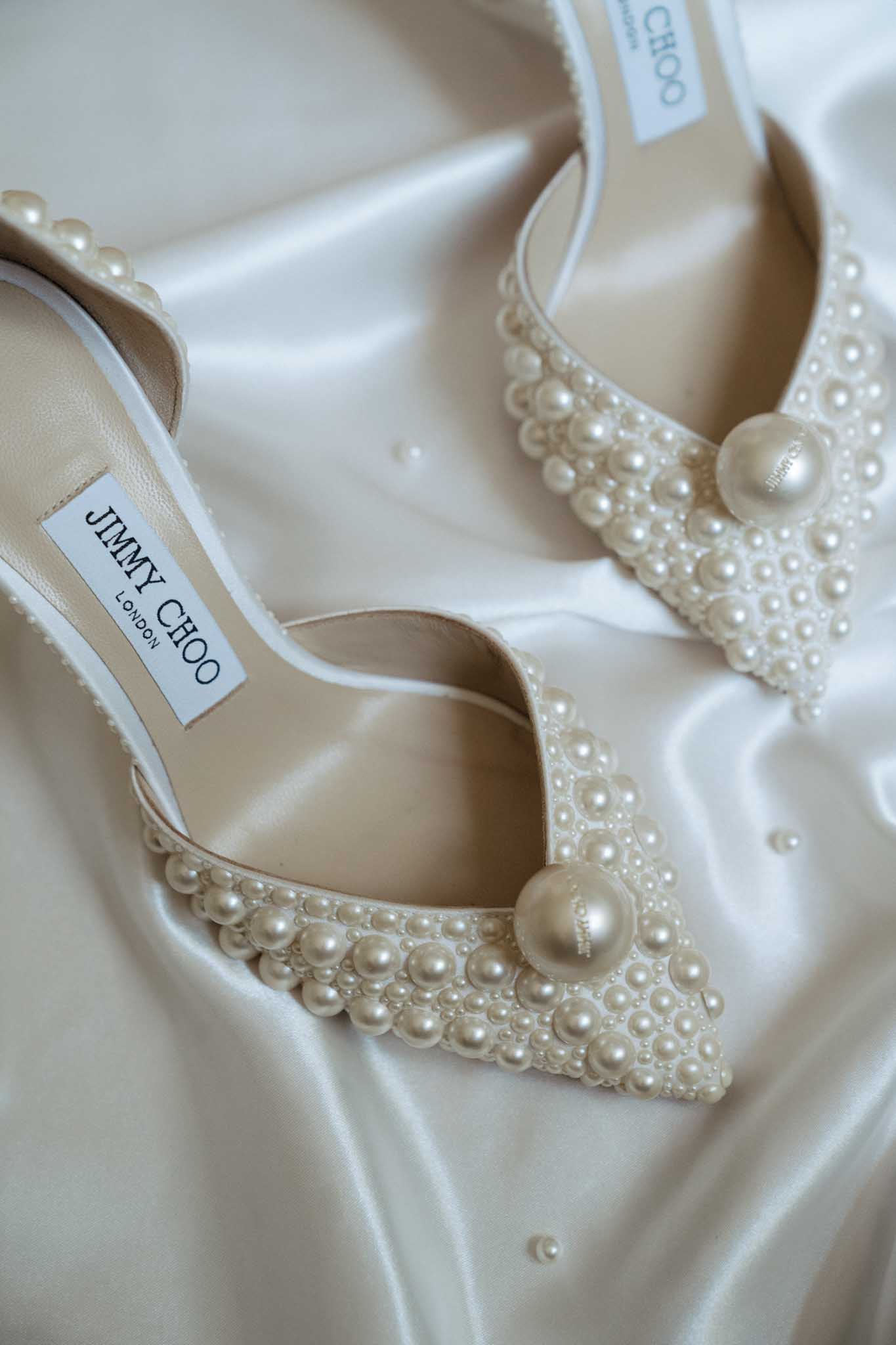 A close-up detail shot of a pair of Jimmy Choo London pointed-toe bridal heels covered in cream and ivory pearl embellishments of varying sizes, each shoe featuring a large central pearl at the toe strap. The shoes are displayed on ivory satin fabric, which creates soft folds and a reflective sheen in the background. The Jimmy Choo branded insole label is visible on the left shoe. The styling is classic bridal with a pearl-forward aesthetic.