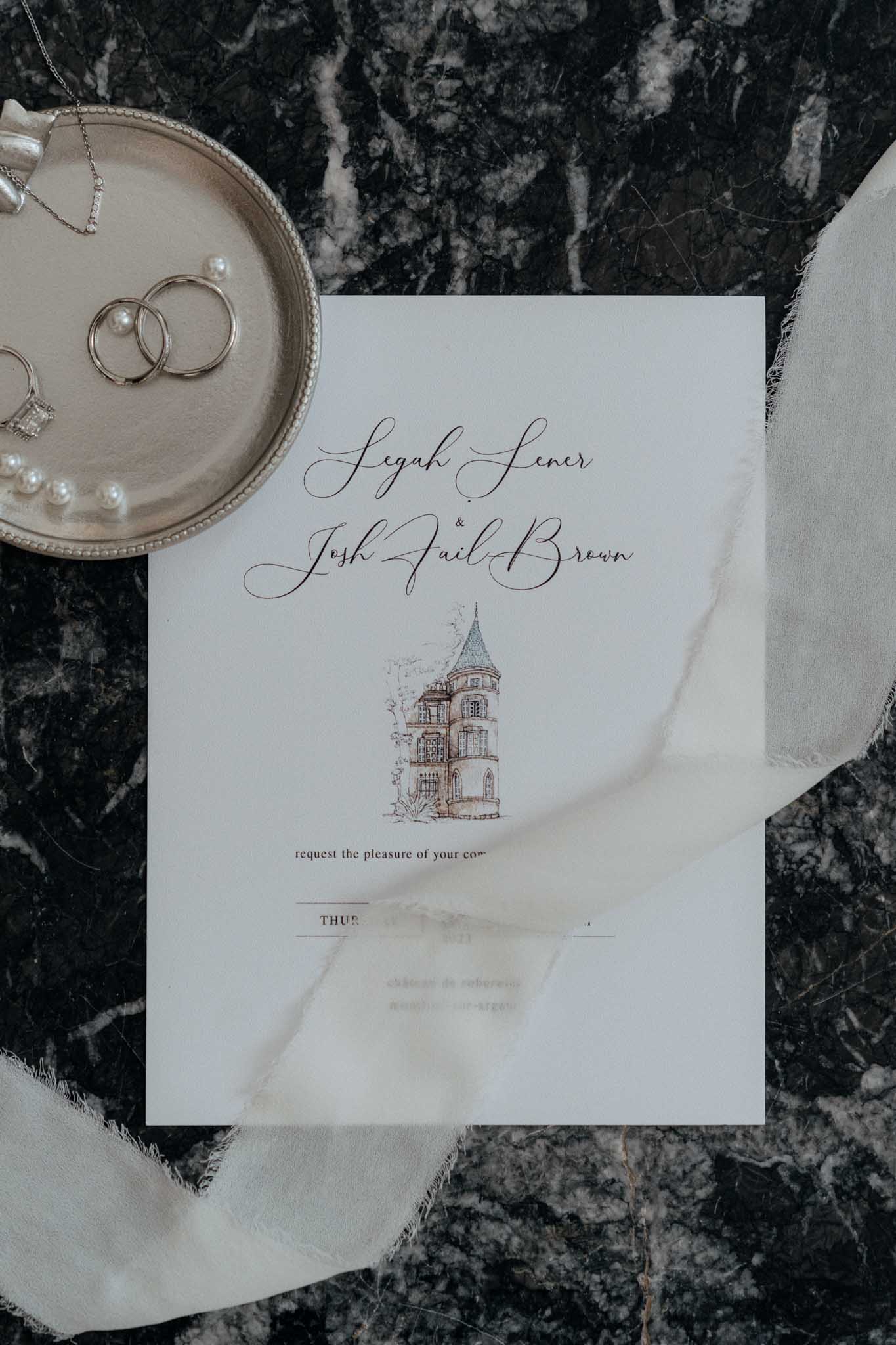 A flat lay detail shot of a wedding invitation suite styled on a dark veined marble surface. The ivory invitation card features the names 'Leah Lener & Josh Tail-Brown' in black script calligraphy, with a hand-drawn watercolor illustration of a French chateau tower in warm brown and grey tones at the center; partially visible text references 'Chateau de Varennes.' The invitation is draped with a frayed-edge ivory silk ribbon. In the upper left, a small silver pearl-rimmed tray holds two gold wedding bands, a diamond engagement ring, a pearl bracelet, and a delicate gold bar necklace.