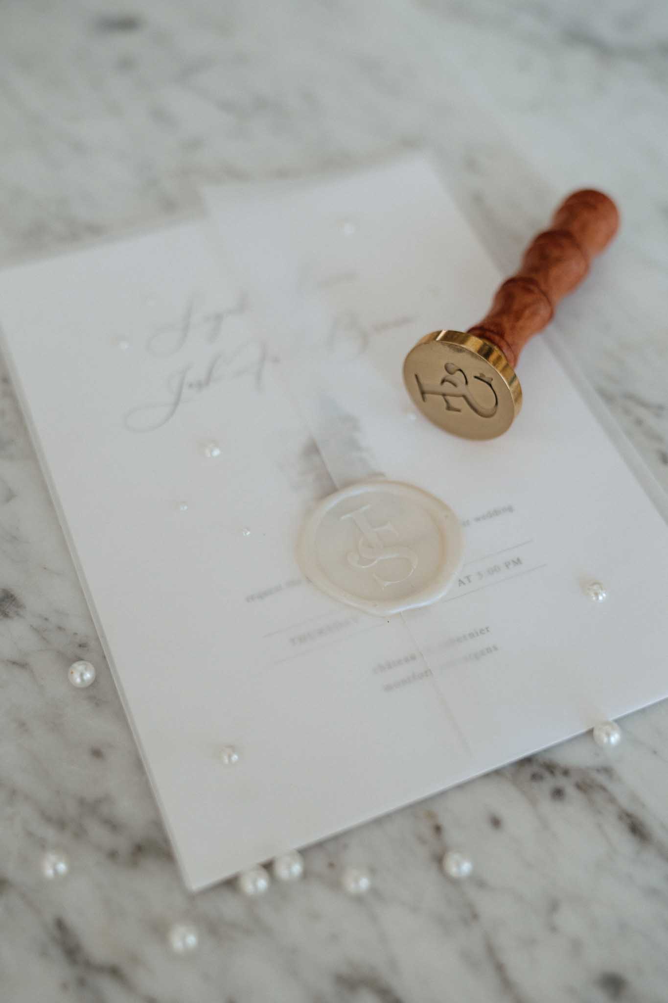 A close-up detail flat lay of a wedding invitation suite in ivory/white, featuring a monogrammed ivory wax seal affixed to the front of the stationery. A brass and wood-handled wax stamp with a matching monogram rests on top of the invitation. The stationery includes calligraphy script for the couple's names and printed event details, and is styled on a marble surface scattered with loose pearl beads. The overall palette is ivory, cream, and gold, consistent with a classic, refined stationery aesthetic.