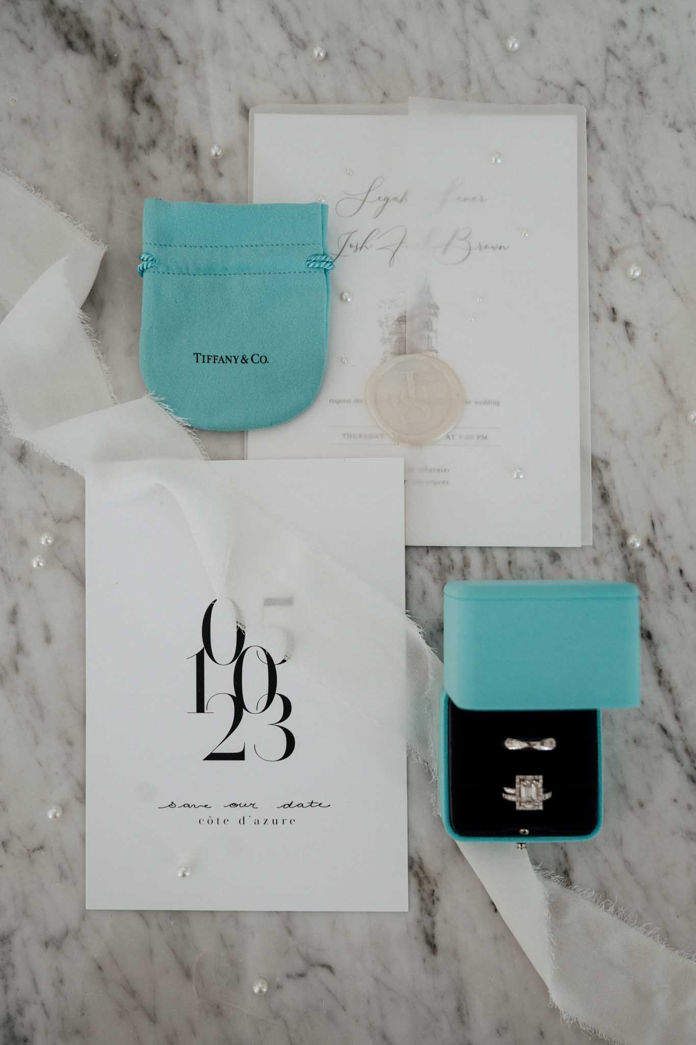 A flat lay detail shot of wedding stationery and jewelry accessories arranged on a white marble surface with scattered loose pearls and ivory silk ribbon. The stationery suite includes a white save-the-date card printed with the date 06/10/23 and the text 'save our date / côte d'azur' in black serif and script typography, alongside a white wedding invitation featuring gold calligraphy script and a château illustration, sealed with a cream wax stamp. A robin's-egg blue Tiffany & Co. velvet pouch sits alongside an open Tiffany blue ring box with a black velvet interior, displaying a diamond engagement ring with an emerald-cut center stone and a matching diamond wedding band, plus what appear to be two additional slim bands. The overall styling palette is Tiffany blue, white, and gold with a clean, modern-classic aesthetic.