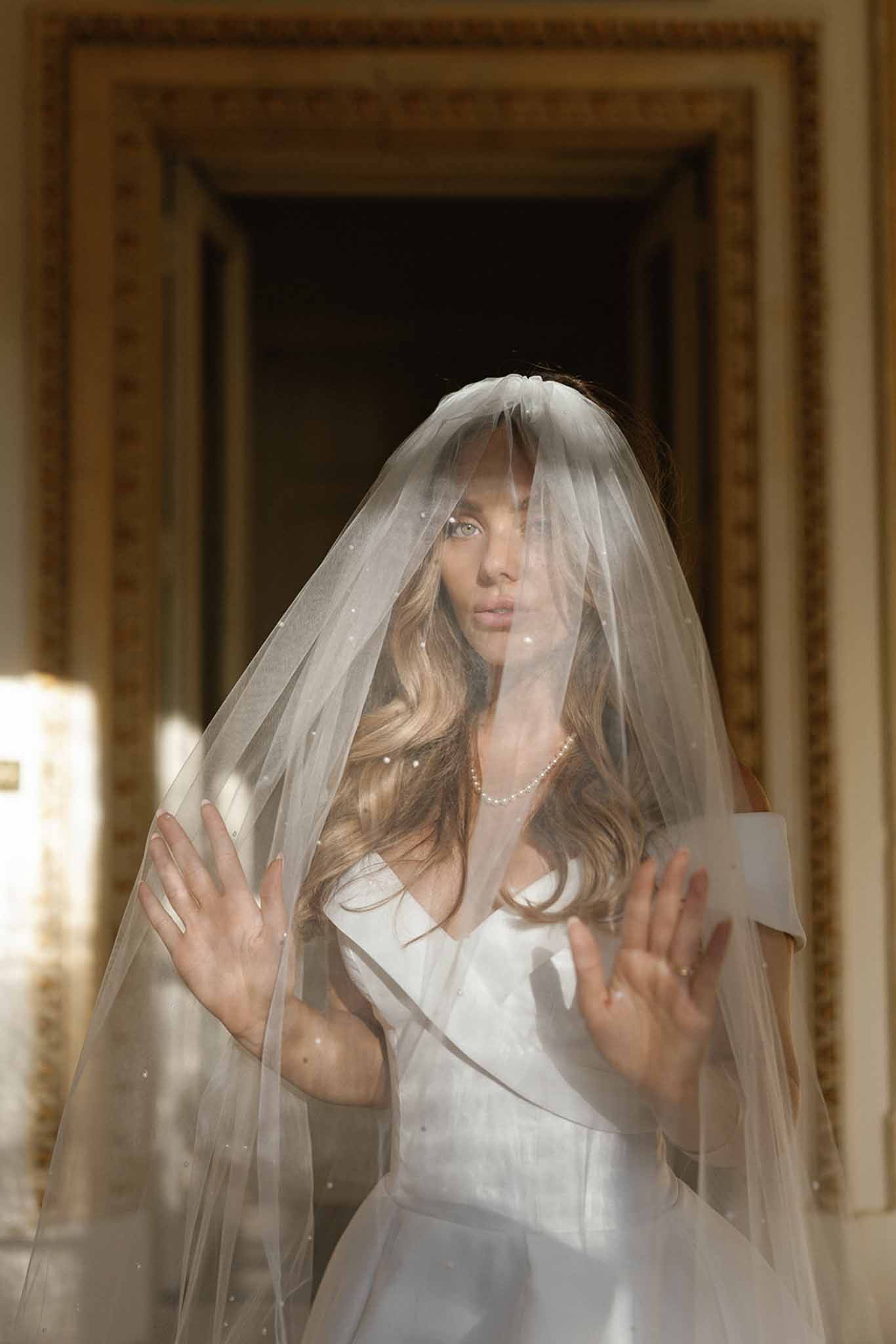 A bridal portrait of a bride standing indoors in front of an ornate gold-trimmed doorframe, shot in natural light with strong directional shadows. The bride wears a white structured strapless gown with a bow or fold detail at the neckline, a pearl necklace, and a floor-length ivory veil scattered with small pearl or crystal embellishments draped over her face and shoulders. Her long, loose wavy blonde hair falls over her shoulders, and she holds both hands up against the veil as if pressing against it. The composition is a close-up portrait framing her from roughly the waist up, with the ornate gold and cream architectural molding of the doorway visible and softly blurred in the background, suggesting a classic French chateau or historic venue interior.