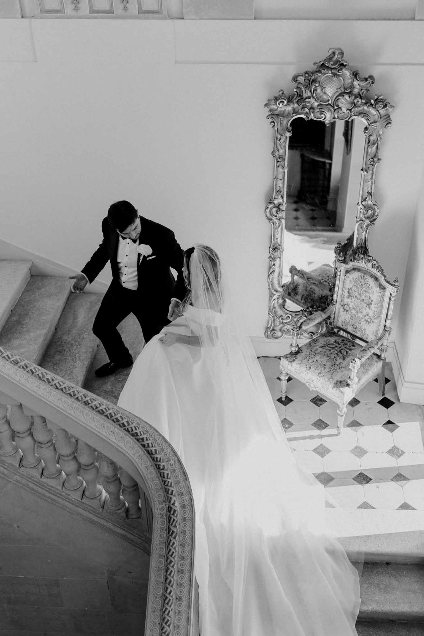 A black-and-white portrait shot taken from an overhead angle, showing a bride and groom on an ornate interior staircase of what appears to be a French chateau or grand historic venue. The groom, dressed in a dark tuxedo with a boutonniere and pocket square, is turned toward the bride and appears to be guiding or reaching for her hand as he descends the stairs. The bride wears a full-length gown with a long flowing veil that spreads across the steps and landing below. The interior features a carved stone balustrade with curved detailing, a geometric marble-inlaid floor, an ornately carved floor-length mirror with baroque-style framing, and a matching antique upholstered chair with carved detailing. The high-contrast B&W tones emphasize the deep shadows of the groom's suit against the bright whites of the bride's veil and gown. Potential venue feature image.