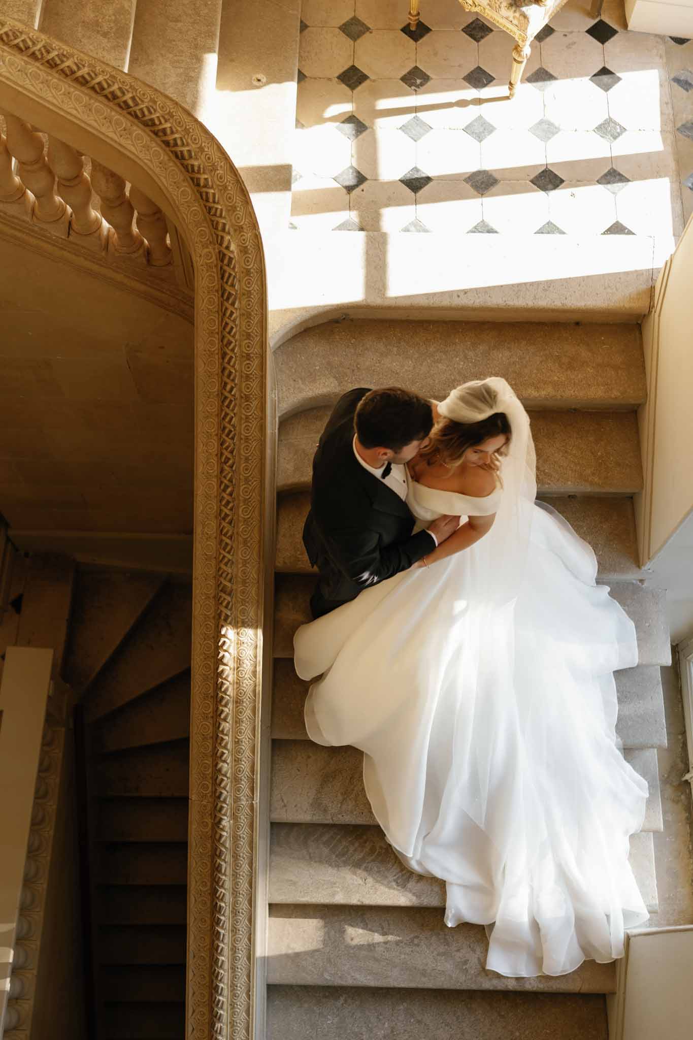 A couple portrait shot from directly above, capturing the bride and groom embracing on a grand interior stone staircase. The groom wears a dark navy or black suit, and the bride wears an off-the-shoulder white ball gown with a voluminous layered skirt and a long veil that cascades across the steps. The staircase features ornate gold-trimmed carved stone archway detailing and a curved balustrade, with black-and-white diamond-patterned floor tiles visible at the top of the frame, indicating a classic French chateau or historic mansion interior. The overhead angle emphasizes the dramatic spread of the dress across the steps, and warm natural light falls across the scene from above. Potential venue feature image.