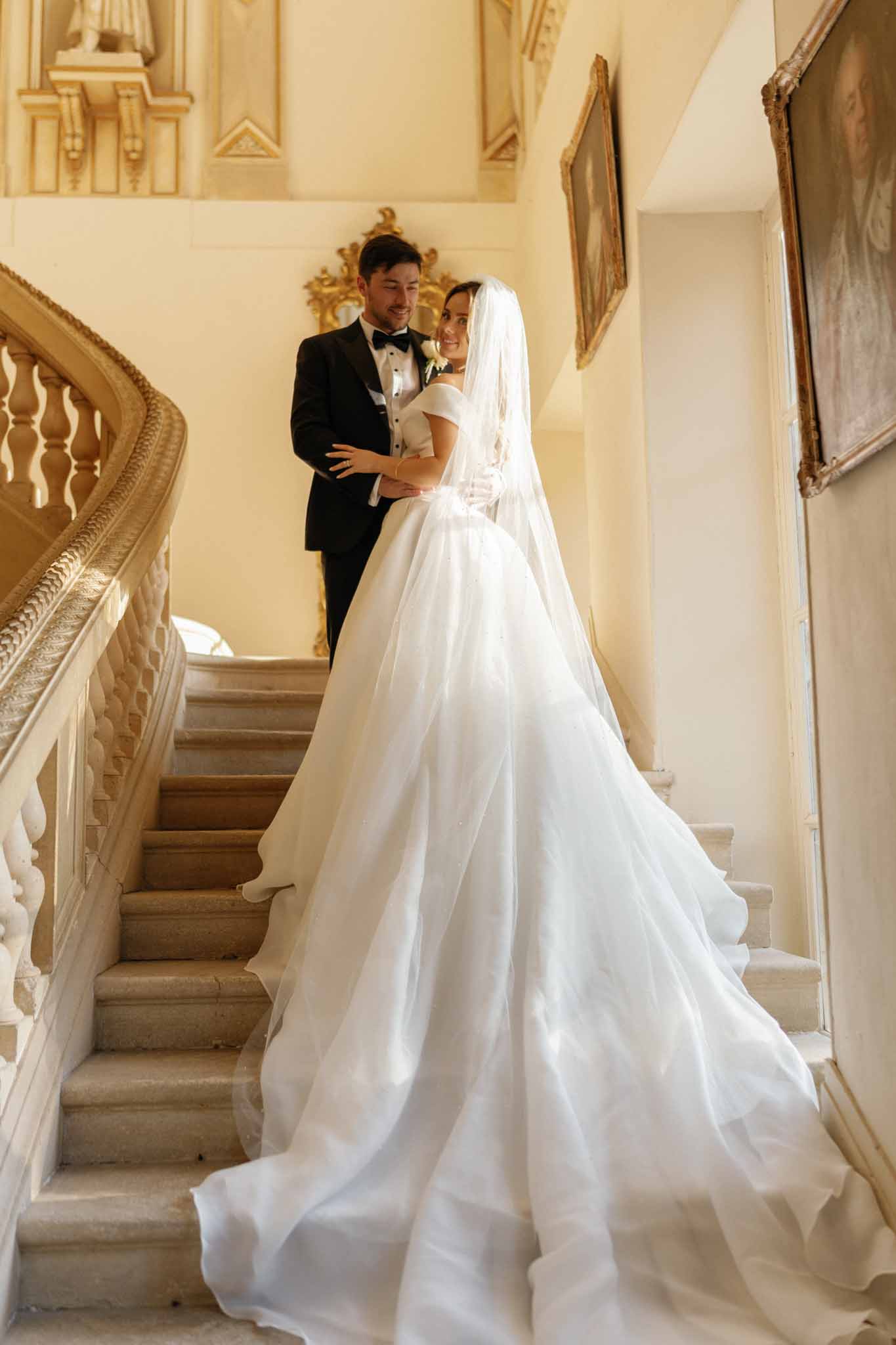 A couple portrait taken on the grand interior staircase of what appears to be a French chateau or historic mansion. The bride wears an off-the-shoulder white ball gown with a voluminous skirt and an extremely long cathedral-length train that cascades down the stone steps, paired with a fingertip-length veil and white gloves; the groom wears a classic black tuxedo with a black bow tie and a single white rose boutonniere. The two stand close together mid-staircase, the groom embracing the bride from behind as she turns to smile at the camera. The interior decor includes gilt-framed oil paintings on warm cream walls, an ornate gilded baroque mirror, and a carved stone balustrade, reflecting a classic formal aesthetic. The shot is a full-length portrait taken from below, emphasizing the dramatic spread of the gown's train filling the foreground of the frame.