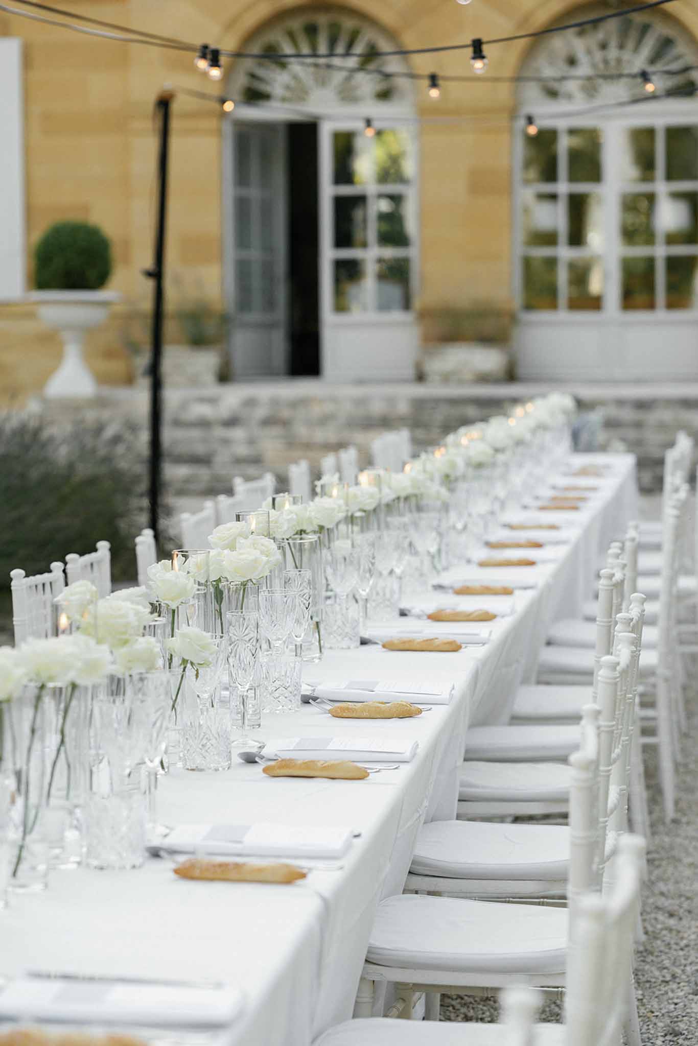 An outdoor wedding reception table setup photographed in a wide portrait-style shot with a shallow depth of field, set in the courtyard of a French chateau with warm yellow stone facade and arched windows visible in the background. The long rectangular dining table is dressed in a white linen tablecloth and lined with white chiavari chairs on both sides. The centerpiece runner consists of white roses and white peonies in clear cut-crystal bud vases of varying heights, interspersed with small votive candles. Each place setting includes white folded napkins, silver cutlery, crystal glassware including tall highball glasses and champagne flutes, and an individual bread roll placed directly on the linen. Overhead Edison bulb string lights are strung above the table, and the overall decor palette is an all-white and clear scheme with a classic, clean aesthetic.