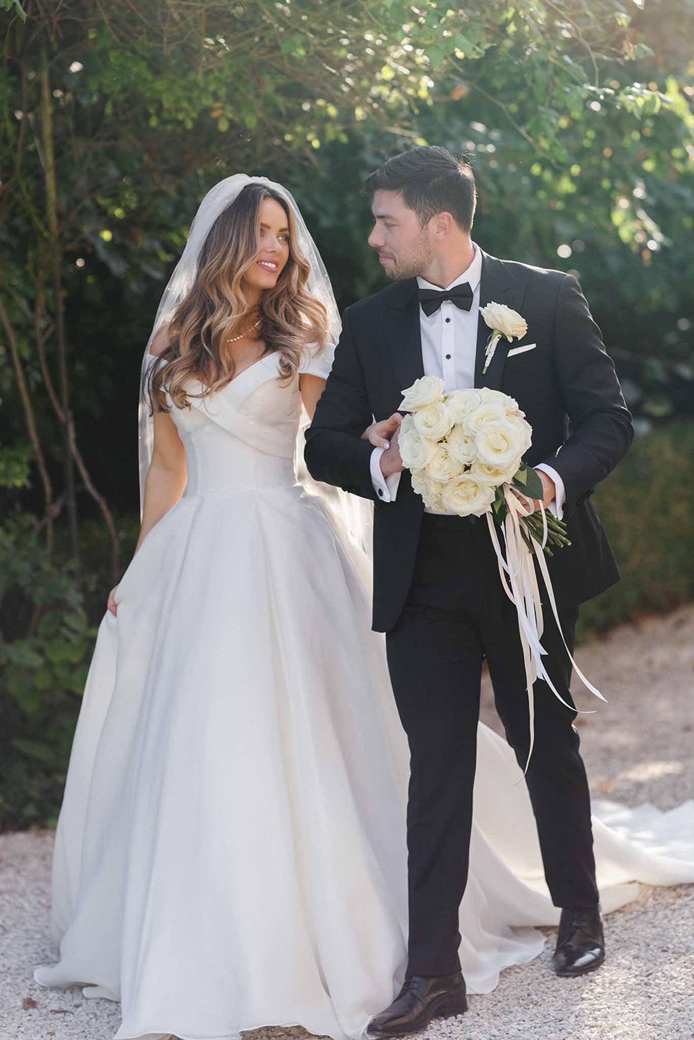A couple portrait taken outdoors on a gravel path with lush green foliage in the background. The bride wears an off-the-shoulder ivory ball gown with a full skirt and cathedral-length train, paired with a shoulder-length veil worn over loose, wavy brunette hair and a delicate gold necklace. The groom is dressed in a classic black tuxedo with a black bow tie, white dress shirt, and a white rose boutonniere. He holds the bride's bouquet, which is a tight round arrangement of cream and white roses with long trailing ivory satin ribbons. The two are looking at each other and smiling as they walk together, captured in a full-length portrait shot with warm natural backlit lighting suggesting late afternoon sun.