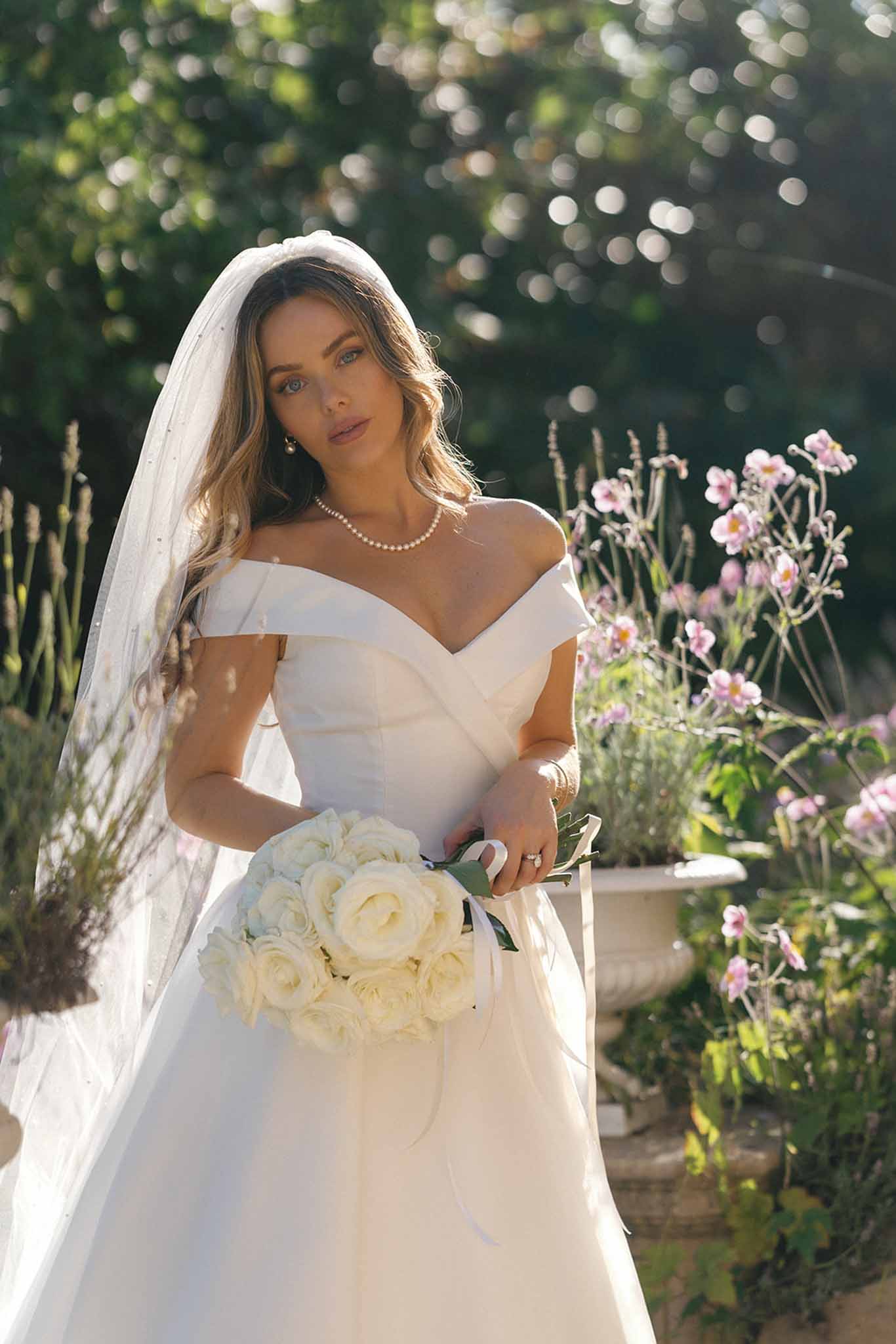 A bridal portrait shot outdoors in a garden setting, featuring a single bride photographed from approximately the waist up. She wears an off-the-shoulder ivory structured gown with a draped sweetheart neckline, paired with a long cathedral-length veil featuring subtle dot detailing. Her accessories include a double-strand pearl necklace and a diamond engagement ring. She holds a tightly gathered round bouquet of cream garden roses with ivory ribbon streamers. Her hair is styled in loose waves, and she wears natural-toned makeup. The background includes a stone planter with pink flowering plants and soft bokeh foliage. The overall styling is classic and polished.