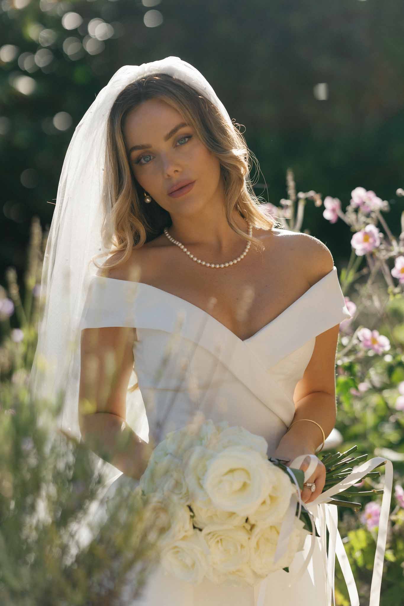 A close-up bridal portrait taken outdoors in a garden setting with soft dappled sunlight. The bride wears an off-the-shoulder ivory structured gown with a draped neckline, a fingertip-length tulle veil, and a pearl necklace and earring set. She holds a bouquet of cream garden roses with ivory satin ribbon streamers and green foliage. Her styling is classic and minimal, with loose wavy hair and natural makeup. Pink blooms are visible in the soft-focus background.
