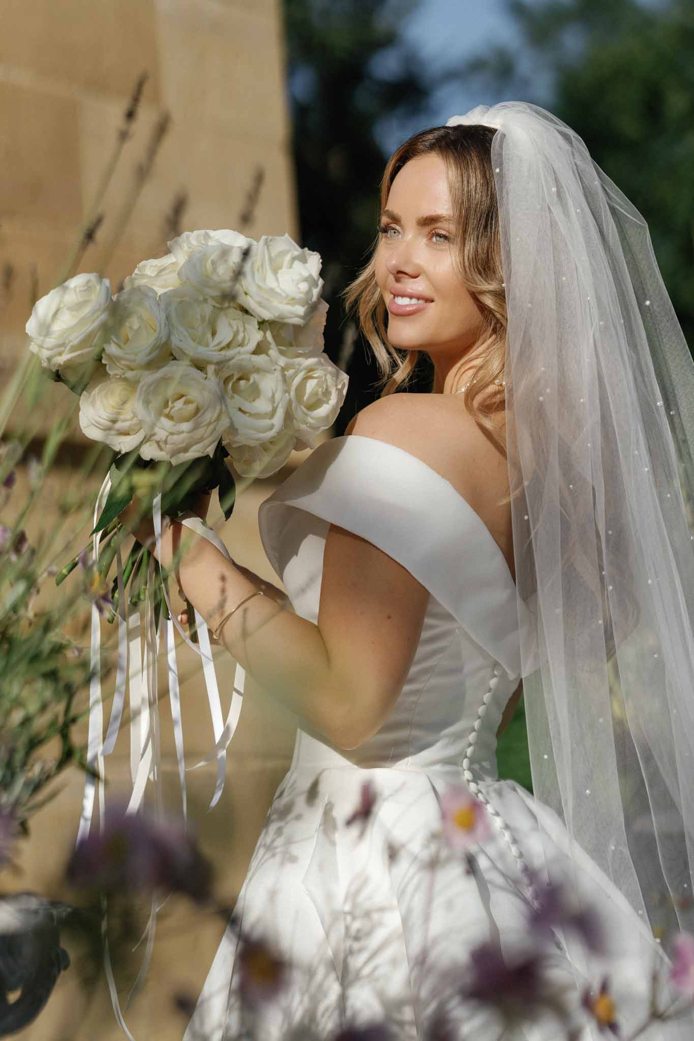 A bridal portrait taken outdoors, showing the bride from approximately the waist up in natural sunlight. She wears an off-the-shoulder white satin gown with button-back detailing and a floral-printed skirt, paired with a pearl-embellished tulle veil. She holds a tightly arranged bouquet of white garden roses with trailing white satin ribbons. The composition is a medium close-up shot from a slightly low angle, with soft foreground blur from wildflowers in muted purple and pink tones framing the bottom of the frame.