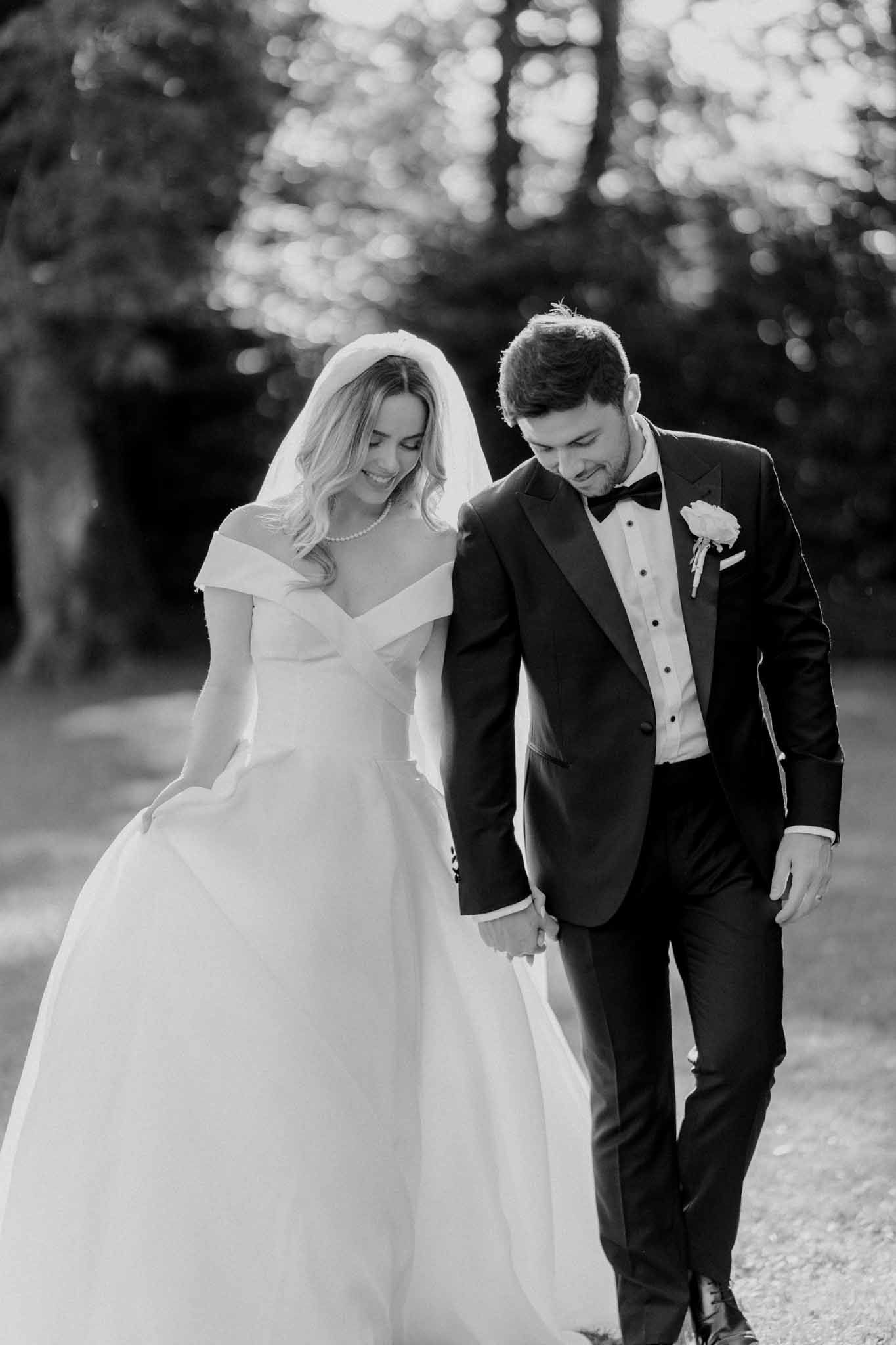 Black-and-white couple portrait taken outdoors, showing the bride and groom walking together and holding hands while both look downward with relaxed smiles. The bride wears an off-the-shoulder ballgown with a crossed bodice, a cathedral-length veil, loose wavy hair, and a pearl necklace. The groom wears a dark tuxedo with a black bow tie, white dress shirt with stud buttons, and a light-toned floral boutonniere on his lapel. The background is heavily blurred with soft bokeh from surrounding trees, creating strong contrast against the bright white of the bride's gown. The image is a mid-length portrait with high contrast and bright highlights on the dress.