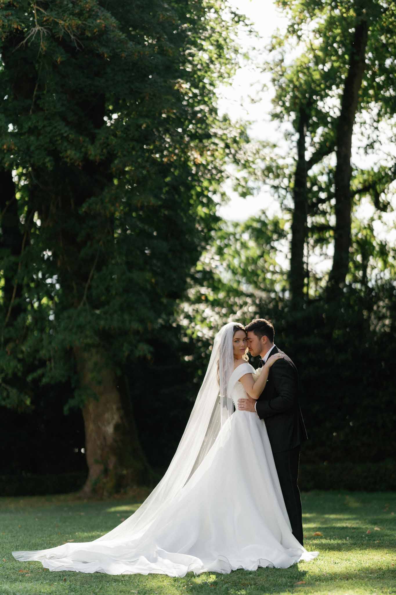 An outdoor couple portrait taken on the grounds of what appears to be a estate or chateau, with the bride and groom embracing in dappled natural light. The bride wears an off-the-shoulder white ball gown with a dramatic cathedral-length train spread across the lawn, paired with a long cathedral veil; the groom is dressed in a classic black tuxedo. The groom nuzzles the bride's temple while she looks toward the camera, in a posed romantic portrait composition. The image is a full-length wide shot with the couple positioned centrally against a backdrop of tall mature trees creating contrast between the dark foliage and the bright white gown.