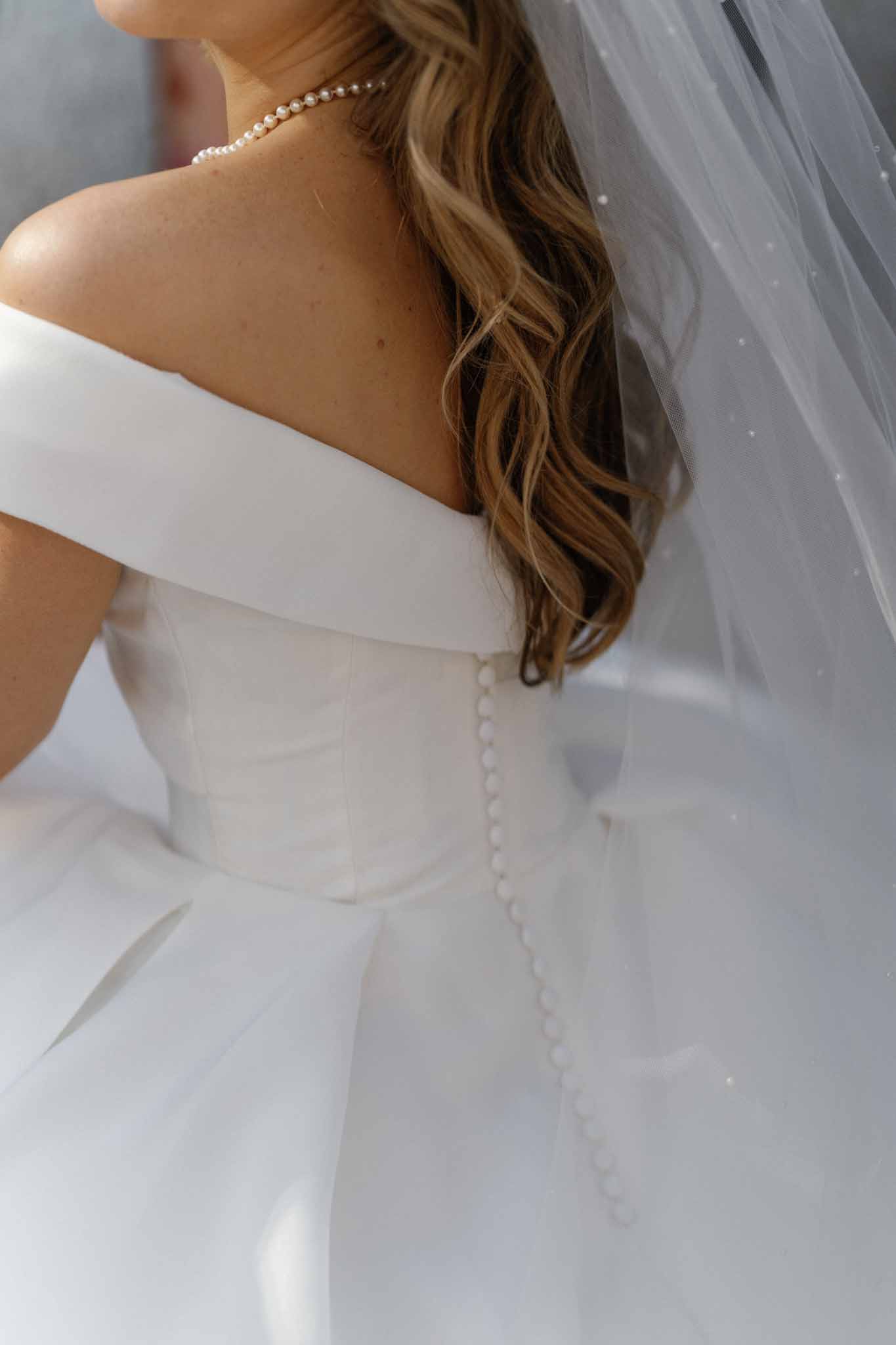 A close-up detail shot of a bride's back during the getting-ready phase, focusing on the construction of her ivory off-the-shoulder ball gown with a structured corseted bodice and a column of small covered buttons running down the center back. A sheer tulle veil with scattered dot embellishments drapes over the full skirt. The bride wears her highlighted brown hair in loose waves, and a classic pearl choker necklace is visible at her neckline. The styling is classic and polished, with a clean, minimal aesthetic.