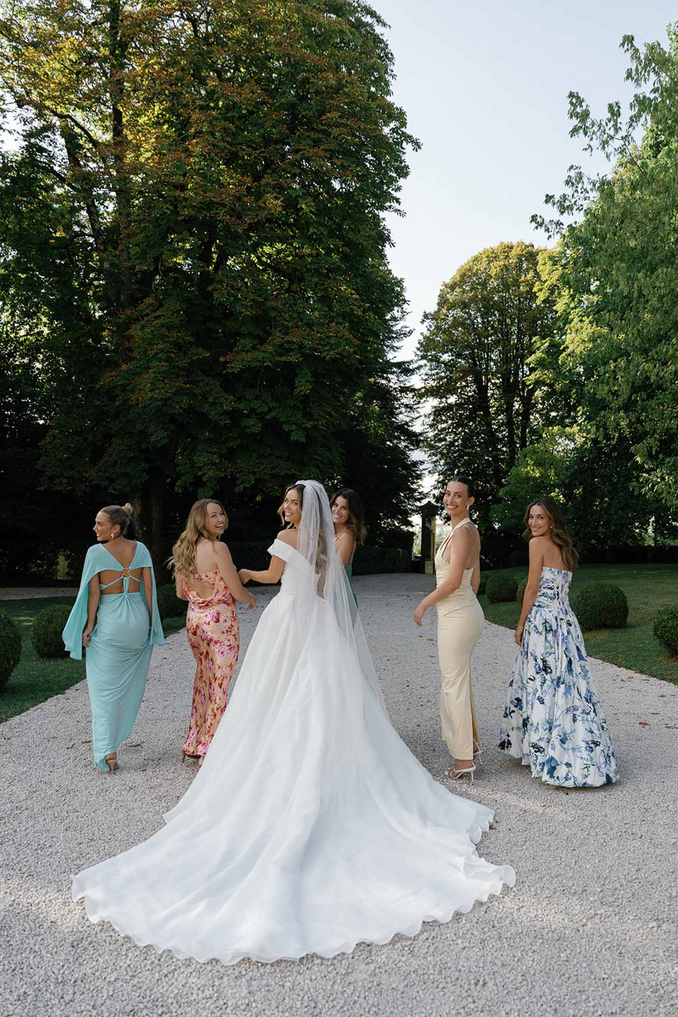 A bridal party portrait taken outdoors on a gravel path in a formal garden, featuring the bride and five bridesmaids or close friends. The bride wears an off-the-shoulder white ballgown with a full cathedral-length train and a long veil, and she is turned slightly toward the camera while looking back over her shoulder. The five women surrounding her wear mismatched formal gowns in a mix of styles and colors: a light aqua blue draped gown with an open back, a pink floral printed satin slip dress, a dark floral dress partially visible, a pale champagne halter-neck column gown, and a white and blue floral printed strapless ballgown. The group is posed walking together on the gravel path, with manicured boxwood hedges and tall mature trees framing the scene in the background, suggesting a chateau or formal estate garden setting. The shot is a full-length wide portrait captured in natural early evening light.